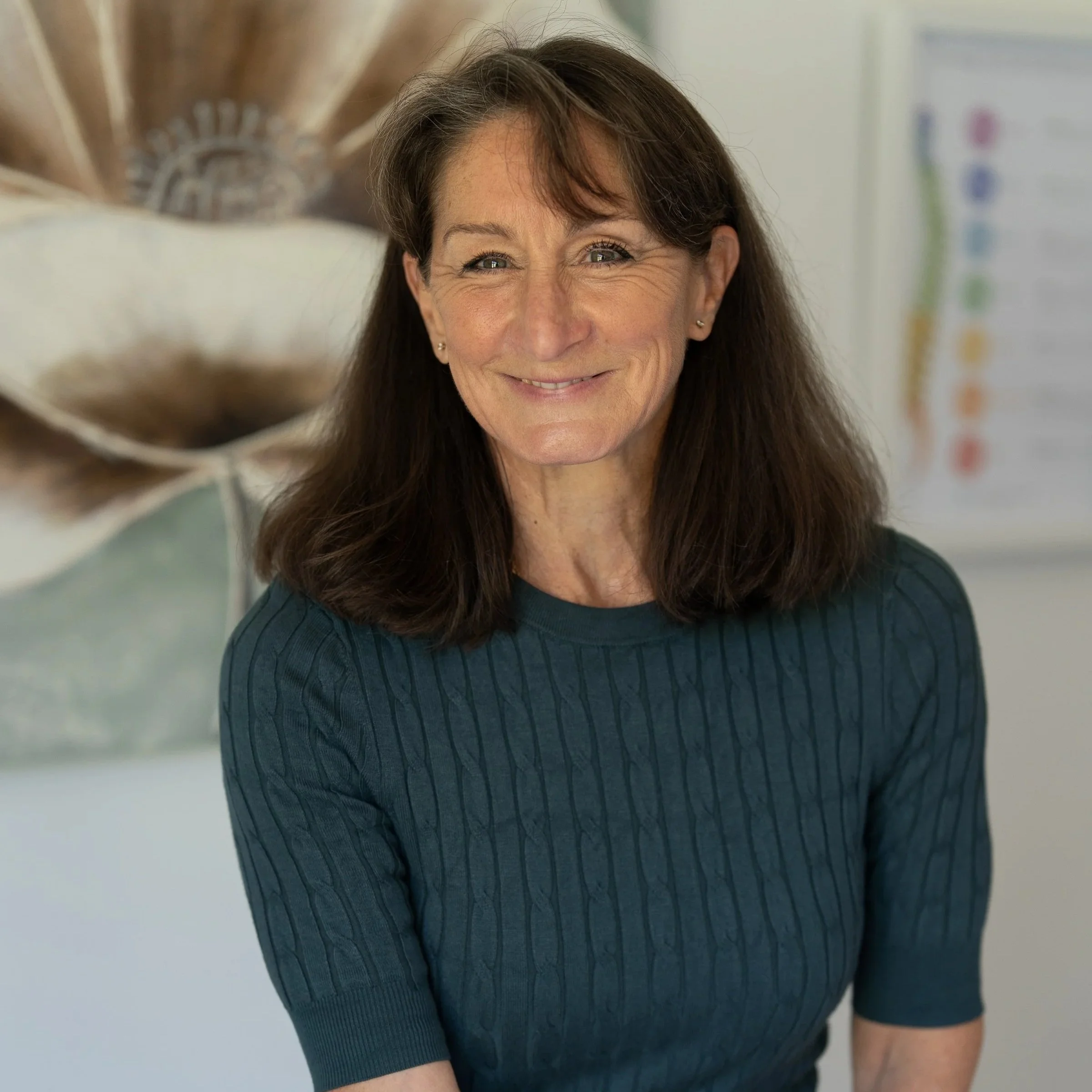 A smiling woman with shoulder-length brown hair wearing a dark green ribbed top, standing indoors with a painting and a chart in the background.