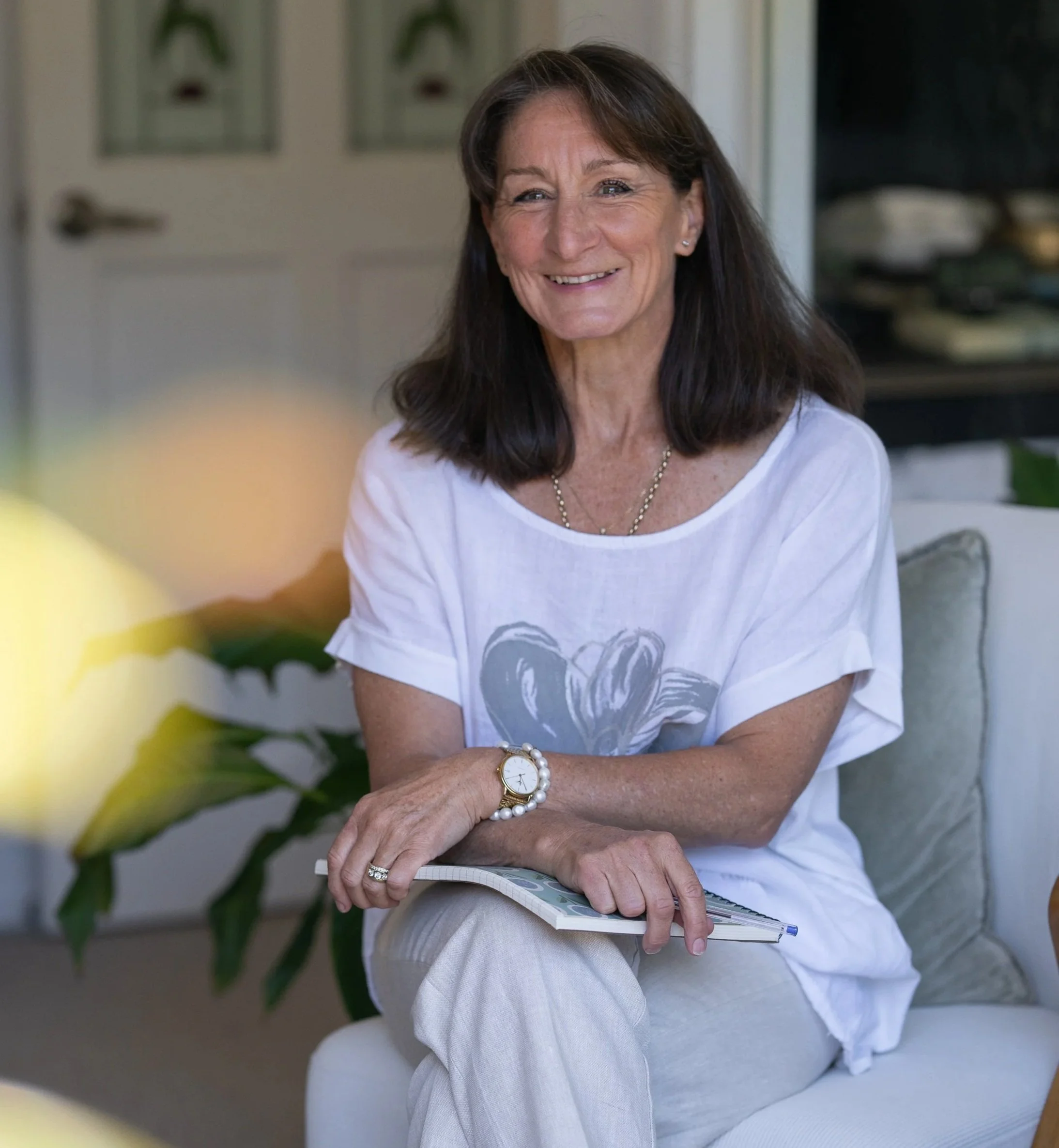 A woman with shoulder-length dark hair and a white t-shirt is sitting on a sofa, smiling, holding a notebook, in a cozy indoor setting with plants and shelves in the background.