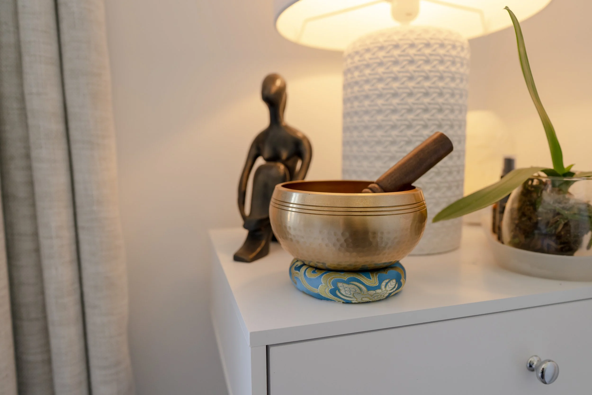 Decorative desktop with a brass singing bowl with a wooden striker, a sculpture of a sitting person, a textured white lamp, and a potted orchid plant.