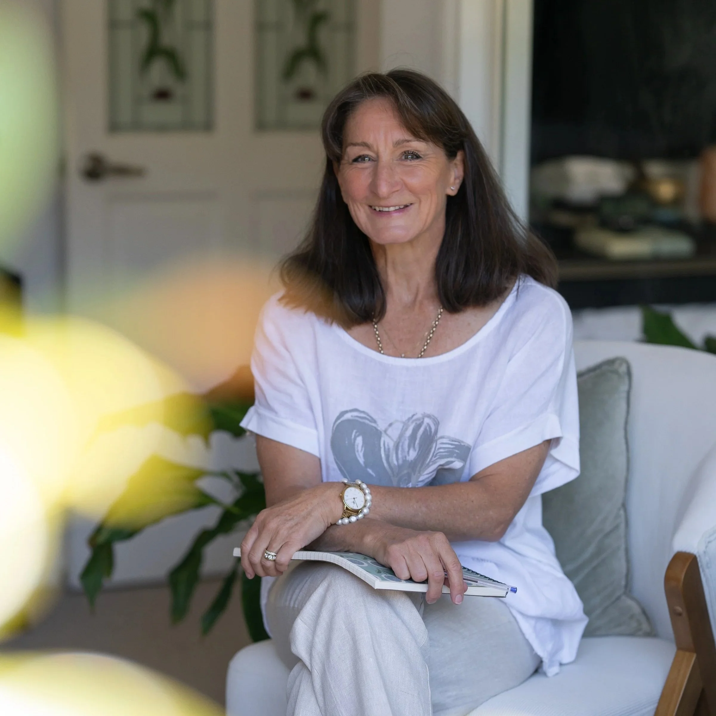 Simone Gilbert - lady with white t-shirt and cream linen pants sitting with legs crossed smiling at camera.