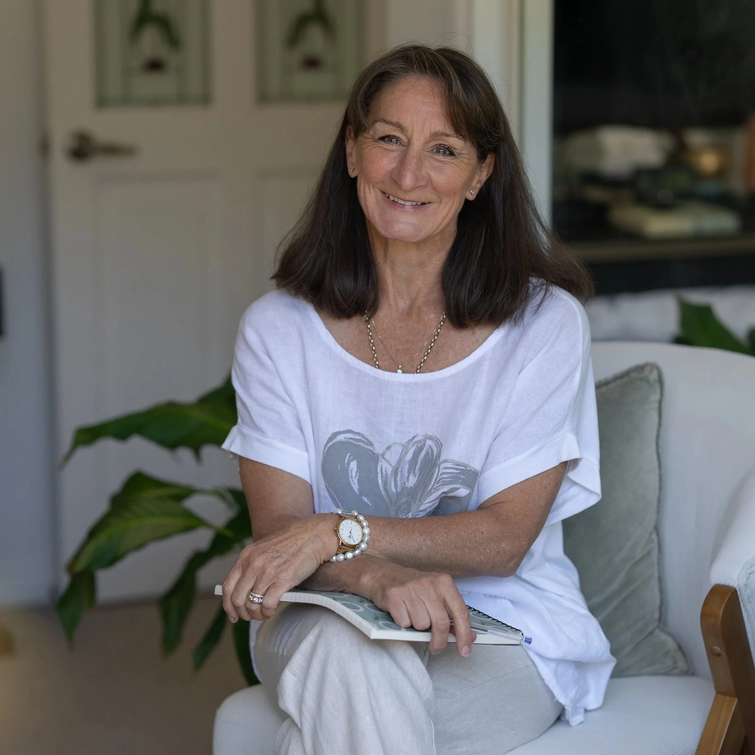 Simone Gilbert - Lady in white t-shirt and linen pants sitting with legs crossed smiling.
