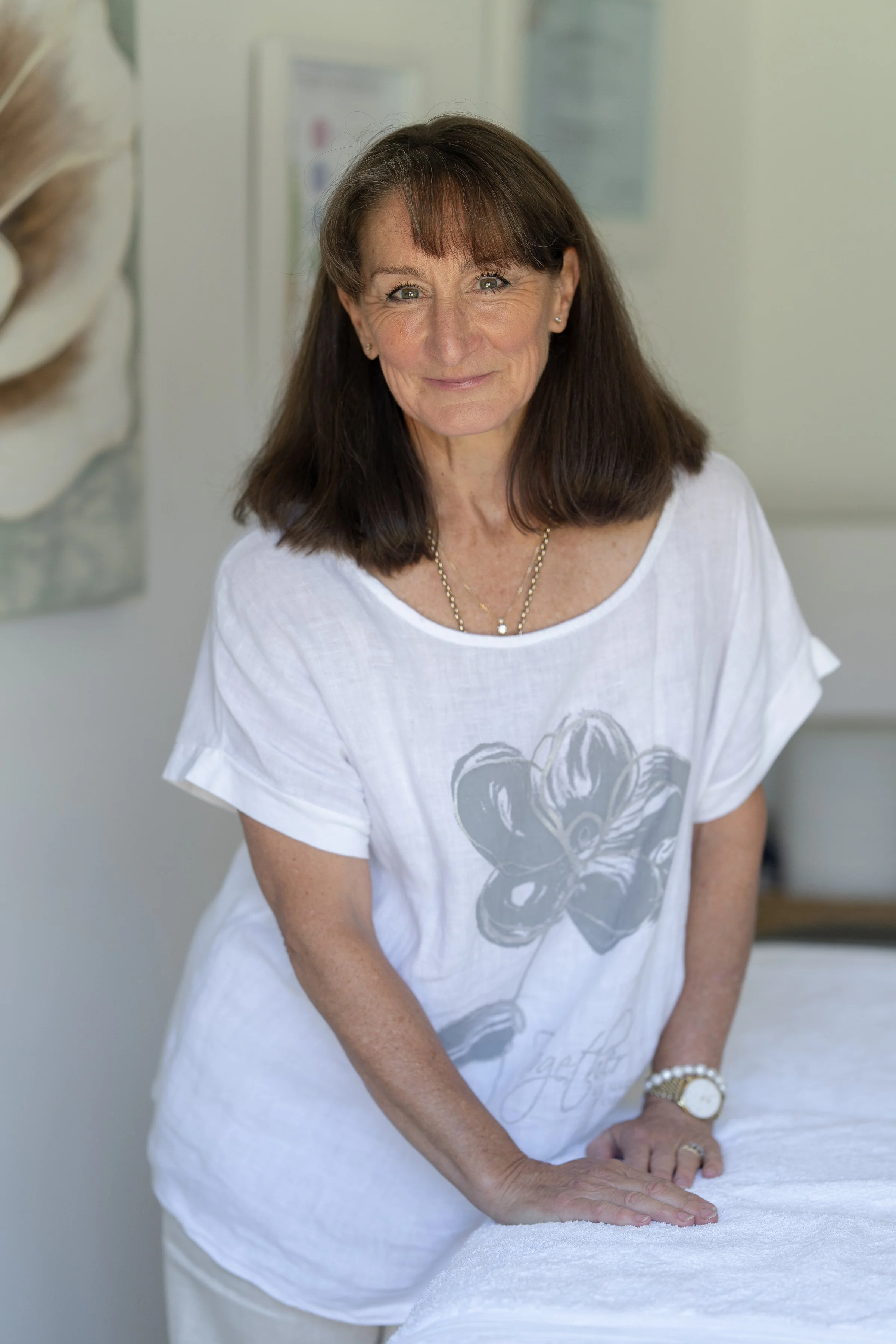 A woman with brown hair and light skin, smiling and leaning on a bed with her hands, wearing a white t-shirt with a large floral design and gold jewelry.