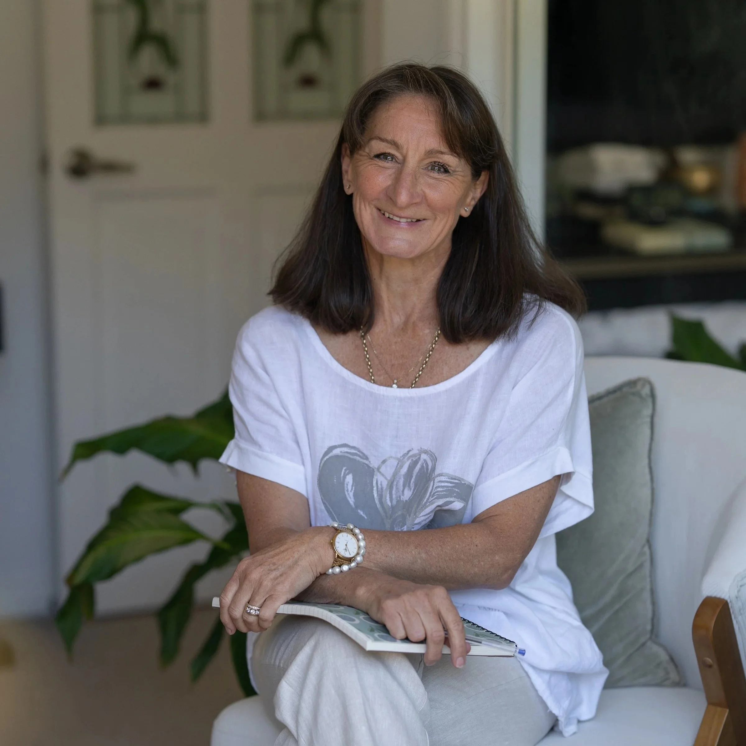 A smiling woman with shoulder-length brown hair wearing a white t-shirt and beige pants sitting on a sofa with a notebook and pen in her lap.