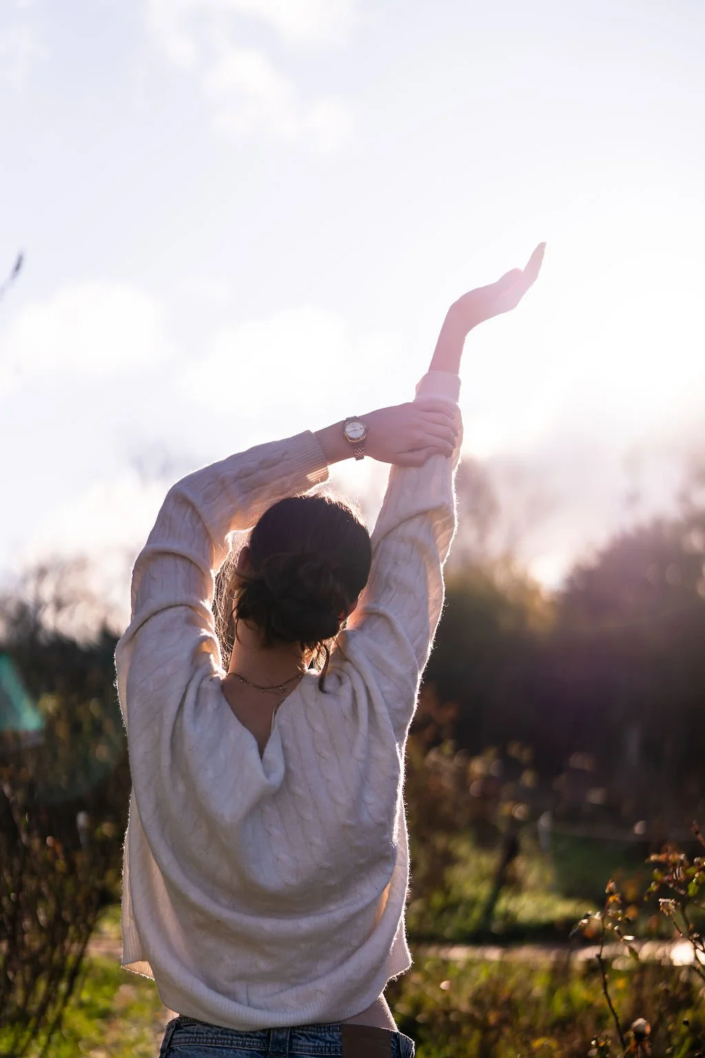 Young lady in garden facing away from the camera with arms up in the air holding onto one arm leaning right.