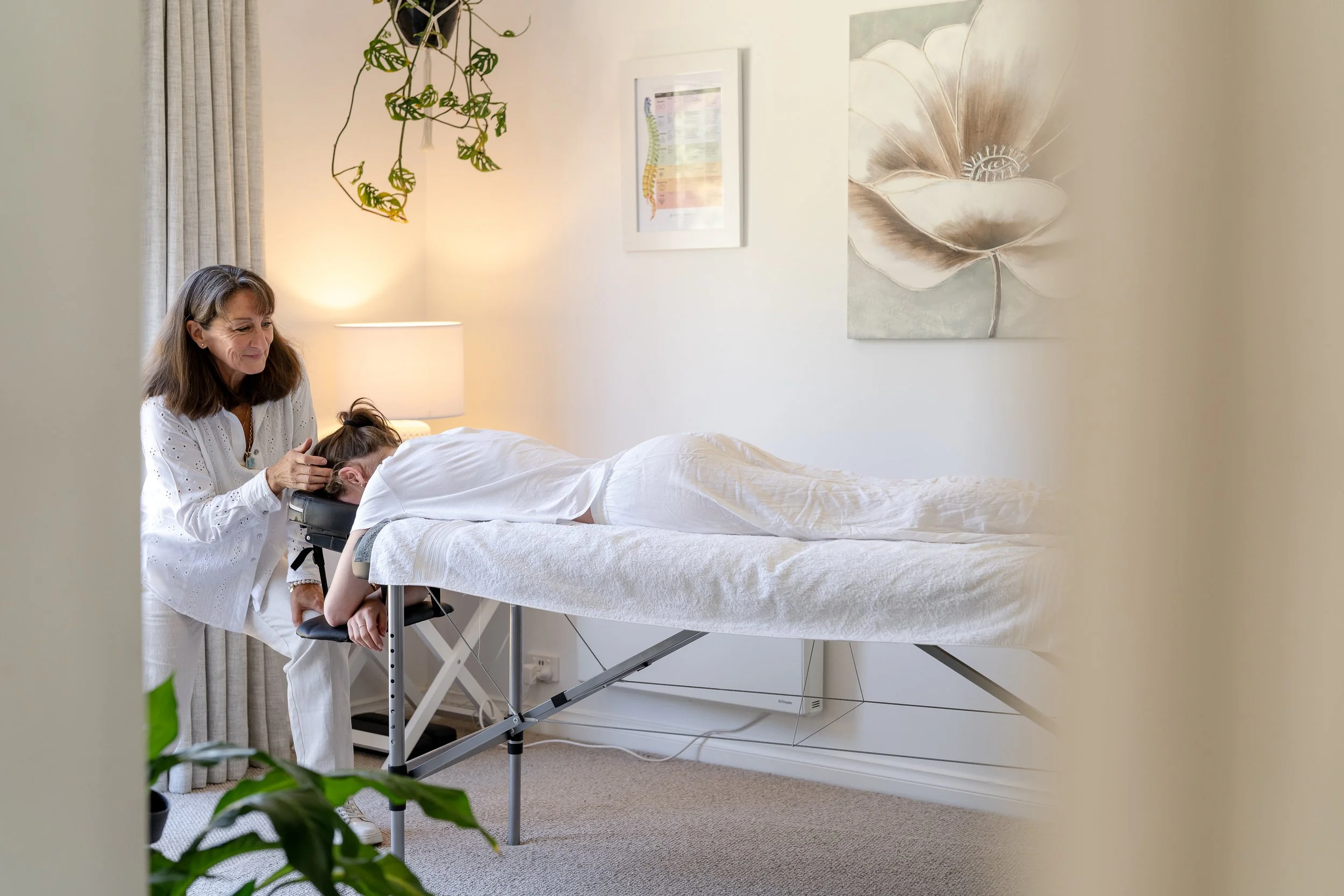 A woman receives Spinal Flow treatment from a therapist in a cozy, well-lit room decorated with artwork and a hanging plant.