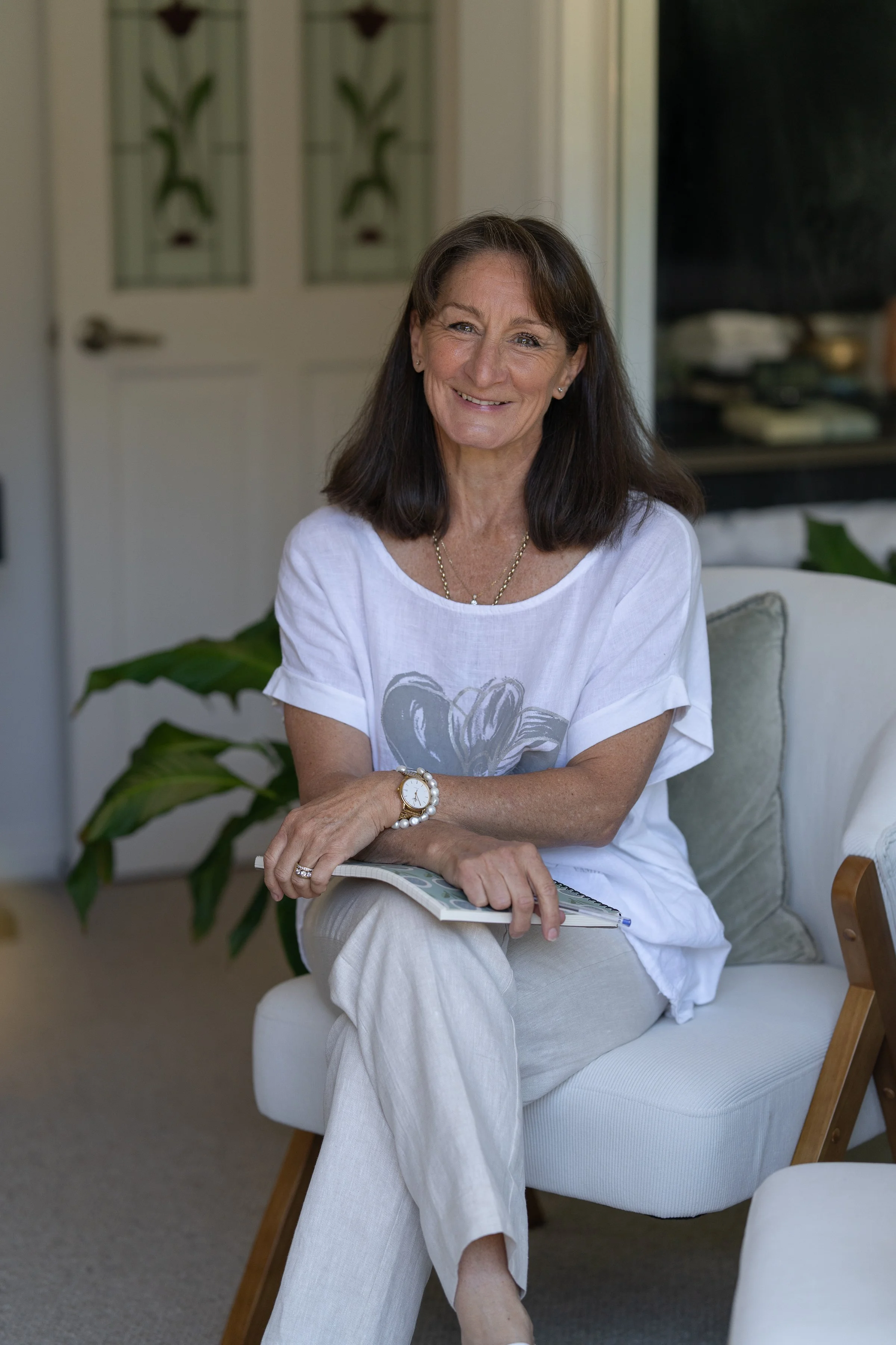 A woman with shoulder-length dark hair sitting on a sofa, smiling, holding a booklet in her lap. She is wearing a white t-shirt and beige pants, with a watch and ring, in a cozy, well-decorated room.