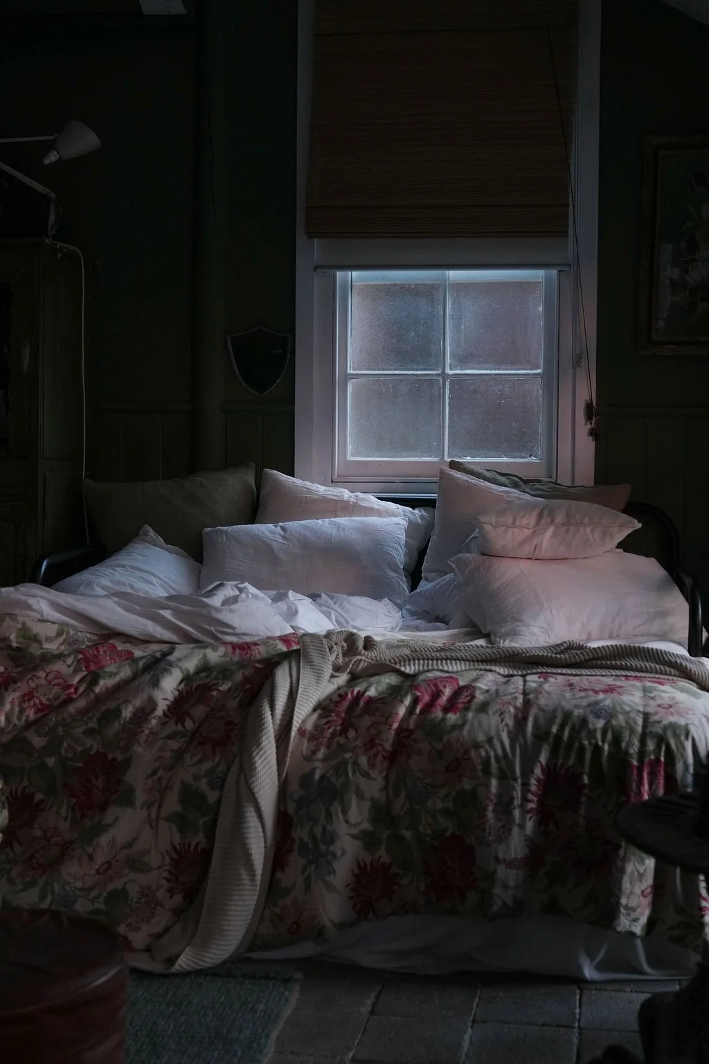 A dimly lit bedroom with an unmade bed, multiple pillows, and a floral bedspread. There is a window with a closed shade, slightly open to reveal frosted glass, and a small nightstand or piece of furniture nearby.