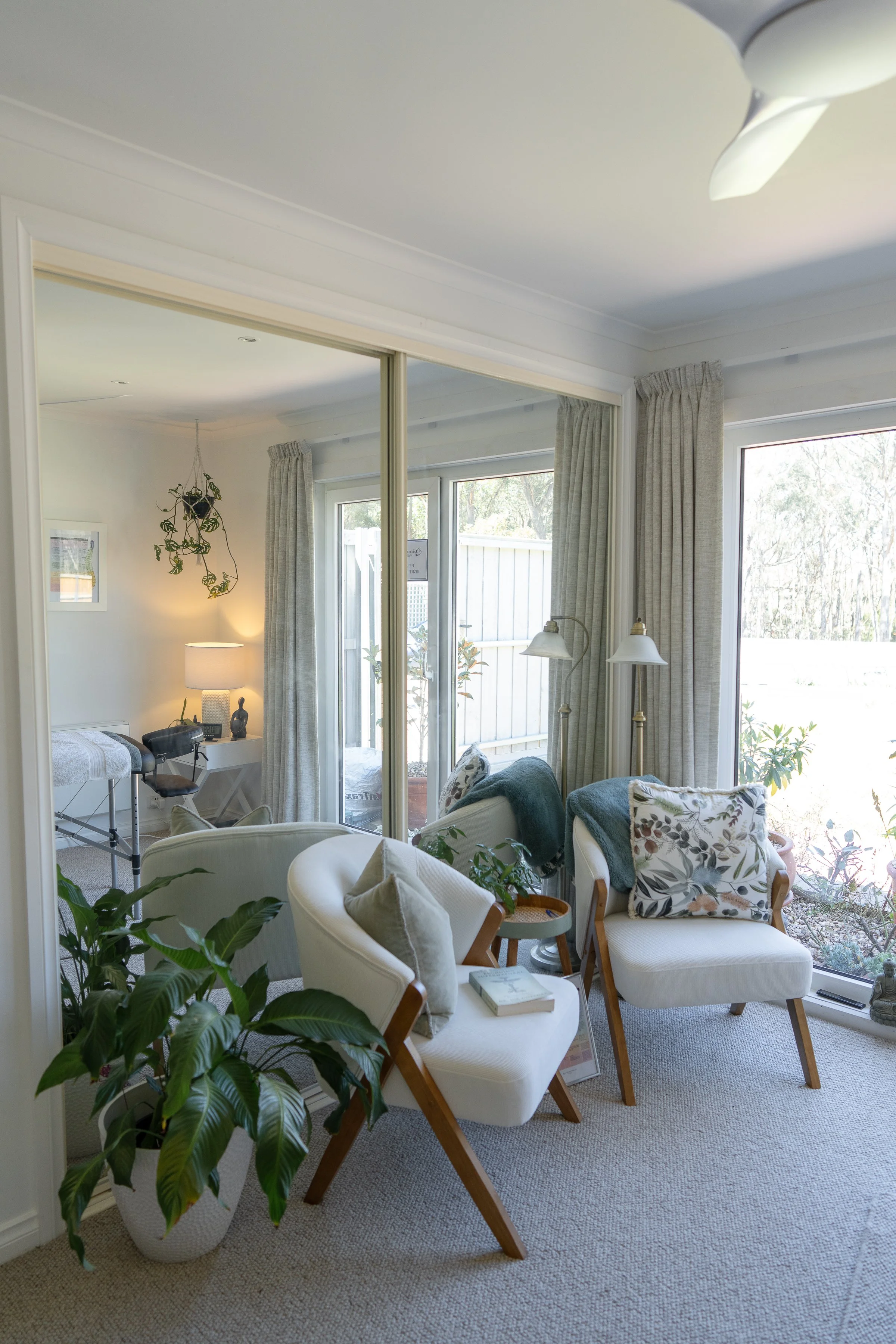 Bright living room with two white chairs, a potted plant, and large windows with curtains, overlooking a garden.