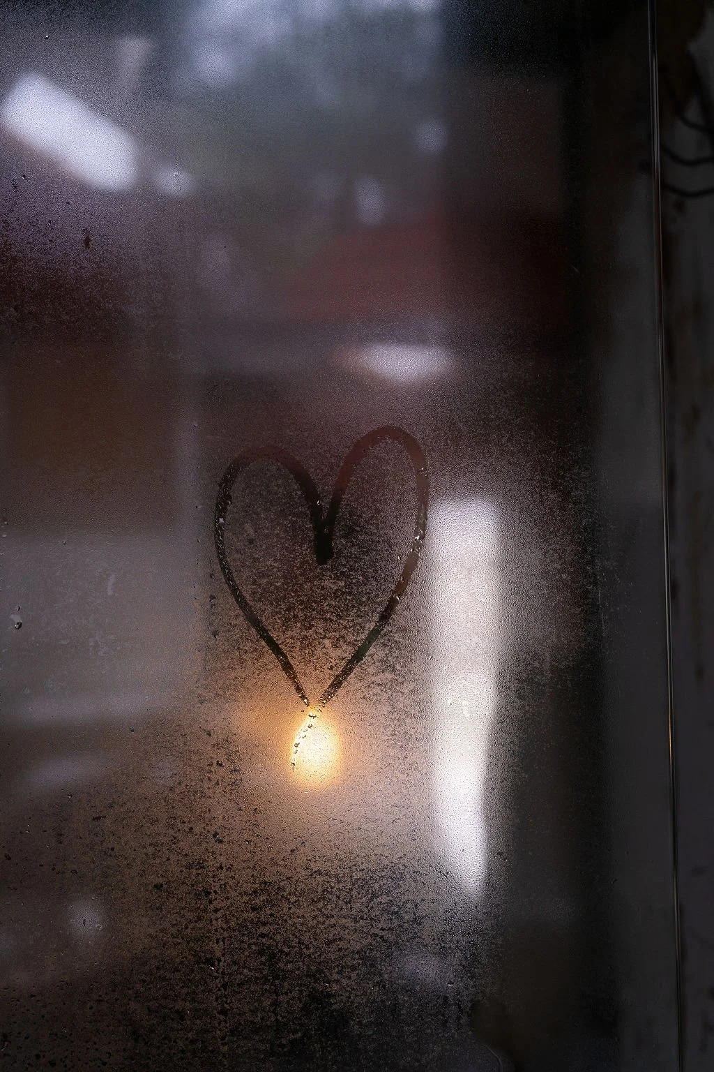 A foggy glass surface with a hand-drawn heart and a small candle or light source beneath it.