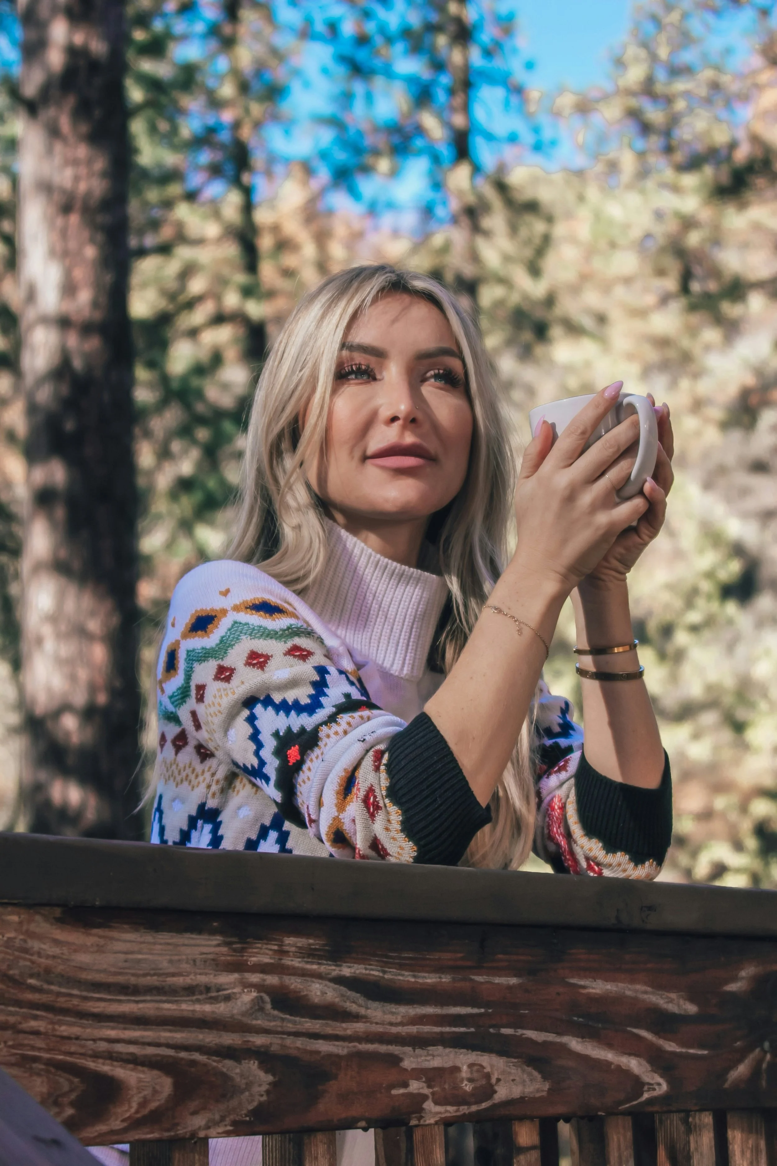 woman in a colorful patterned sweater holds a mug on a wooden porch, surrounded by sunlit forest in a serene outdoor moment.