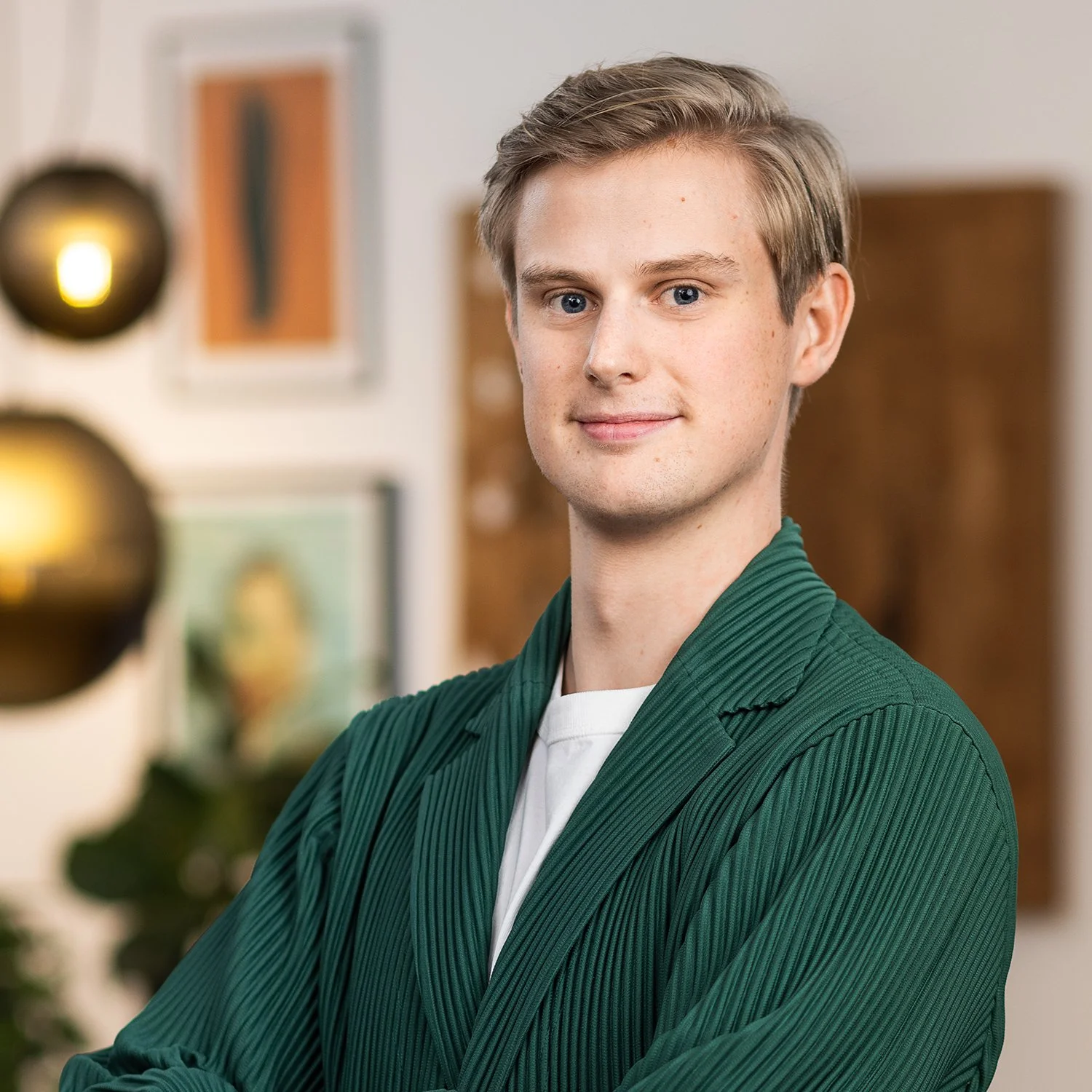 Portrait of a young man with blonde hair and blue eyes, wearing a green jacket and a white shirt, smiling slightly in an indoor setting.