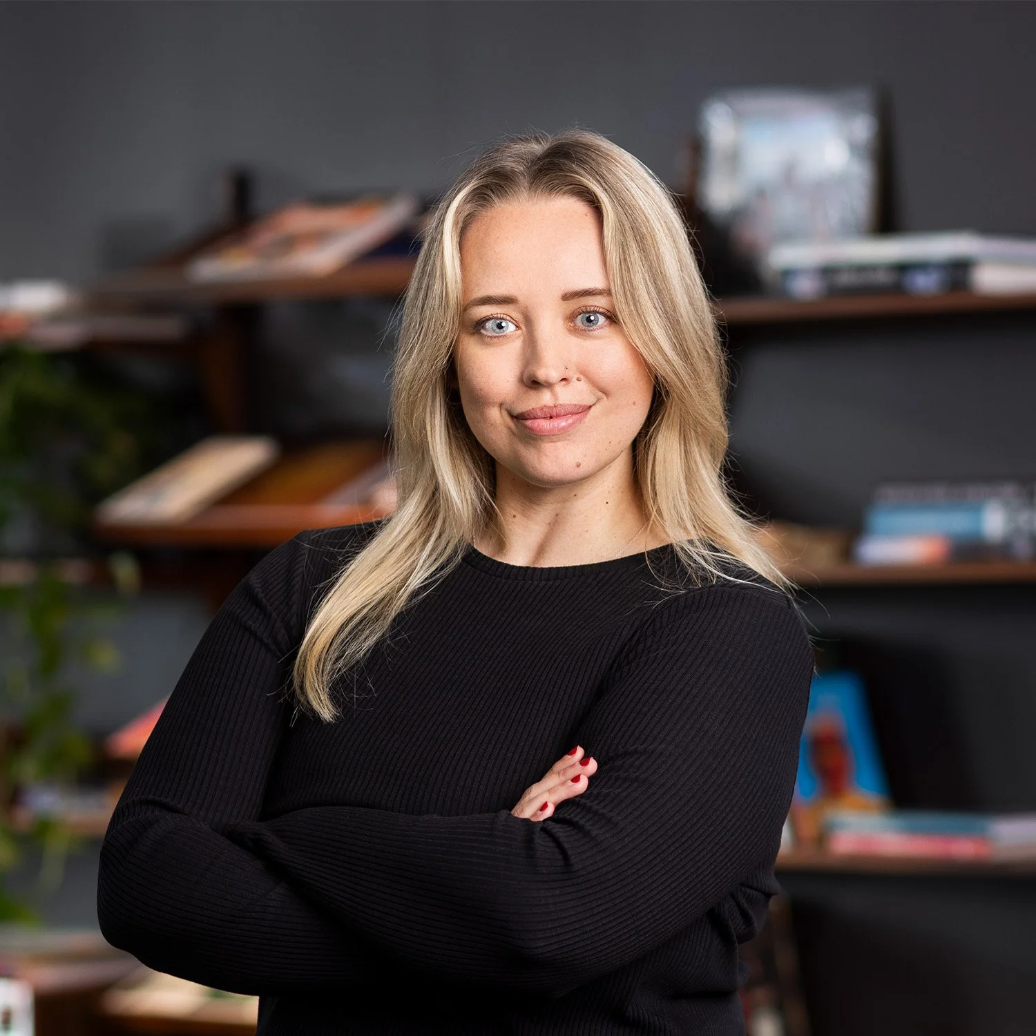 A young woman with blonde hair and blue eyes, smiling, standing with arms crossed in front of a bookshelf filled with books and photos, wearing a black long-sleeve top.