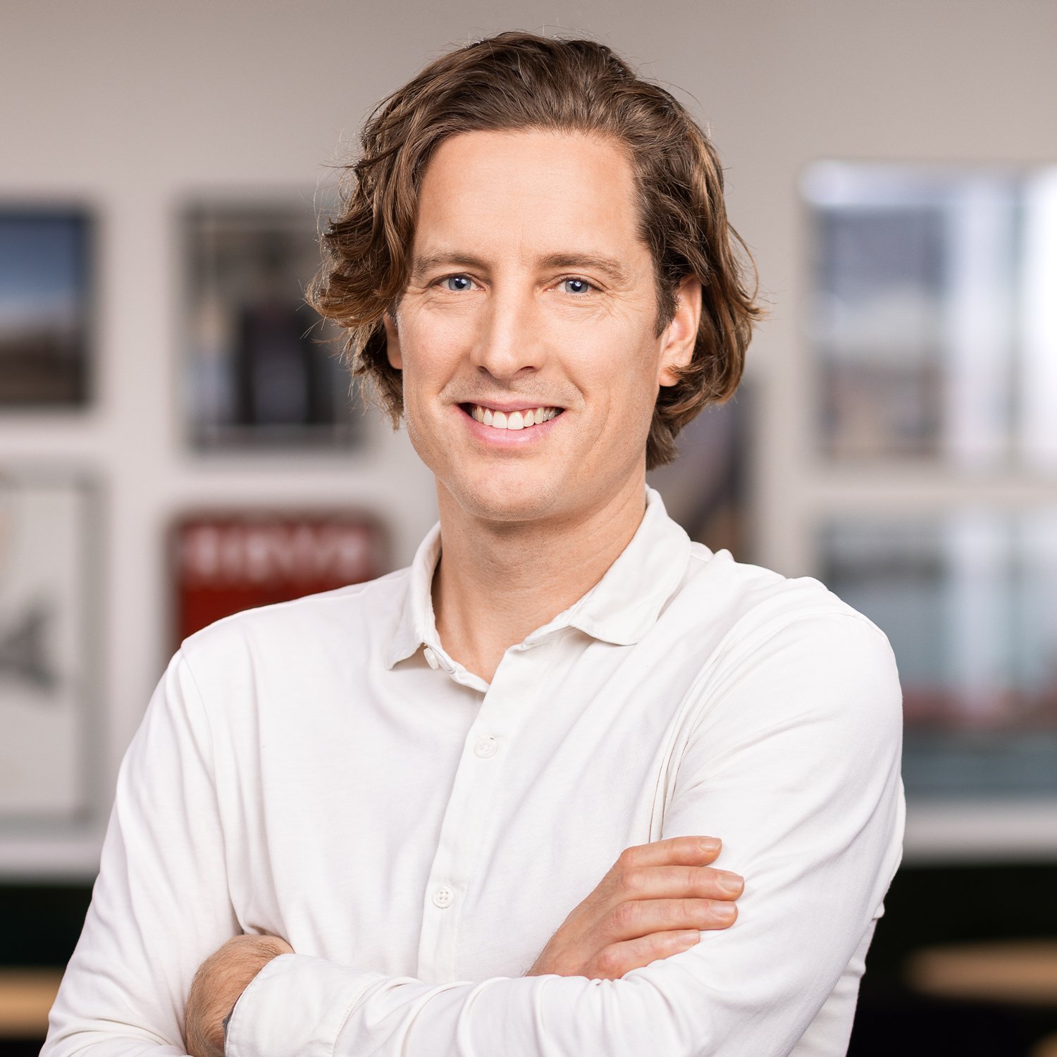 A smiling man with wavy brown hair wearing a white button-up shirt, crossing his arms.
