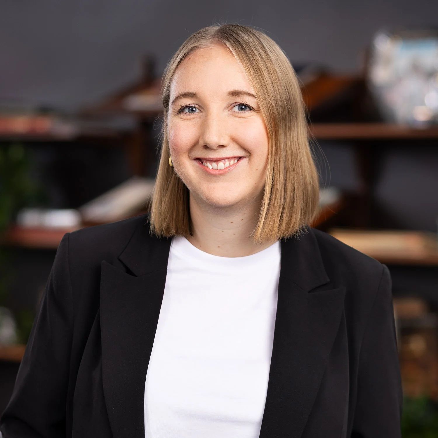 Portrait of a smiling woman with shoulder-length blonde hair, wearing a black blazer and white top, standing indoors with blurred background.