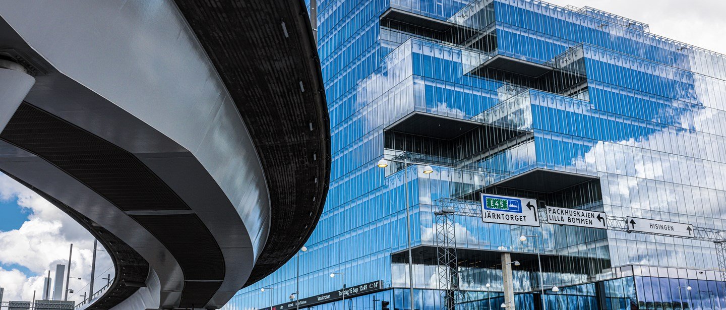 Modern glass building with reflective windows, blue sky, and white clouds, along with an overpass and road signs indicating directions to various locations.
