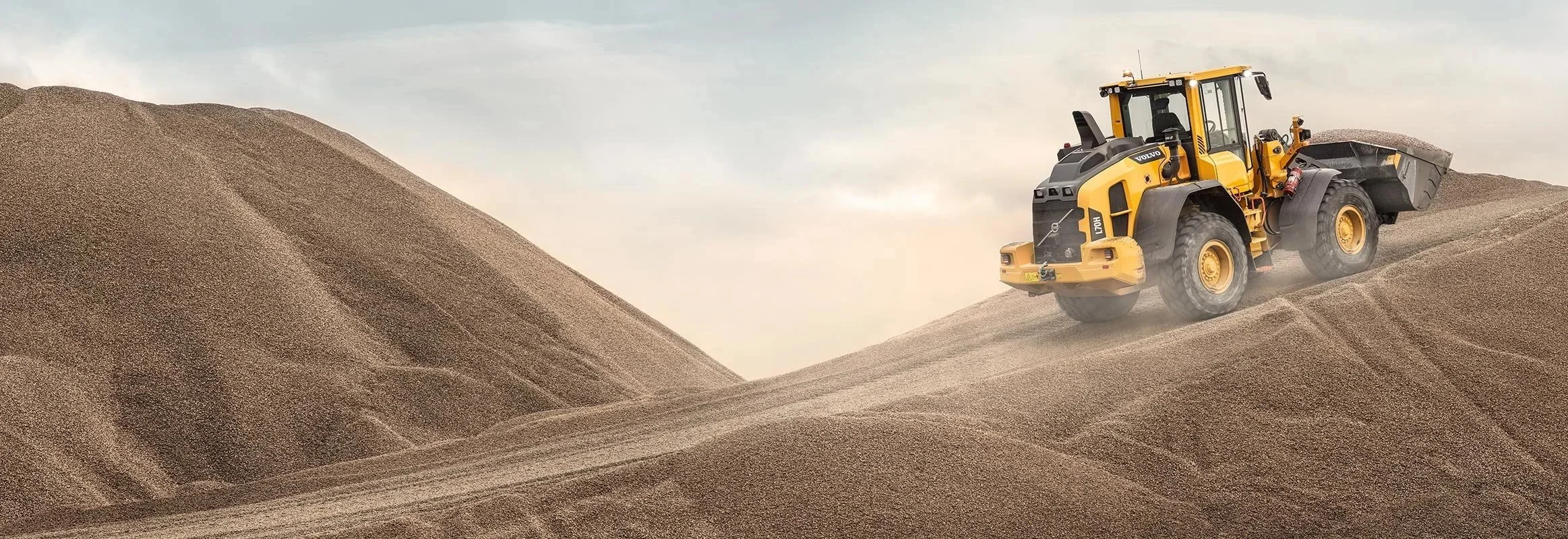 A yellow and black Volvo wheel loader scooping sand on a large sandy hill under a cloudy sky.