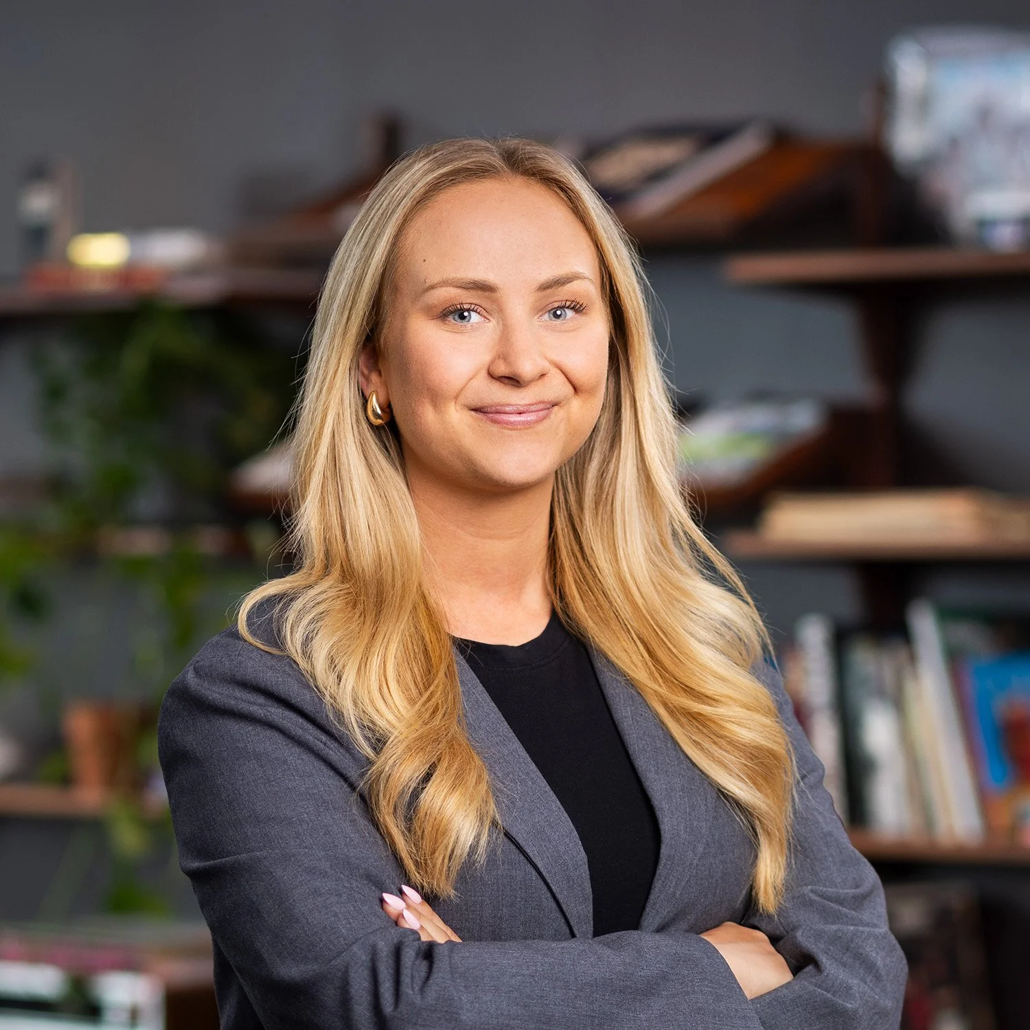 A professional woman with blonde hair and blue eyes smiling with arms crossed, wearing a grey blazer and black top, standing in an office or library setting.