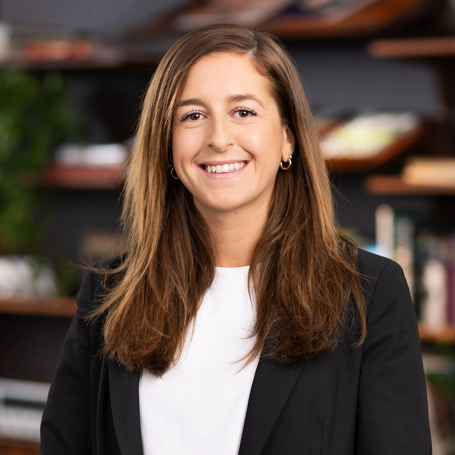 A smiling woman with long brown hair, wearing a black blazer and white top, standing indoors with bookshelves in the background.
