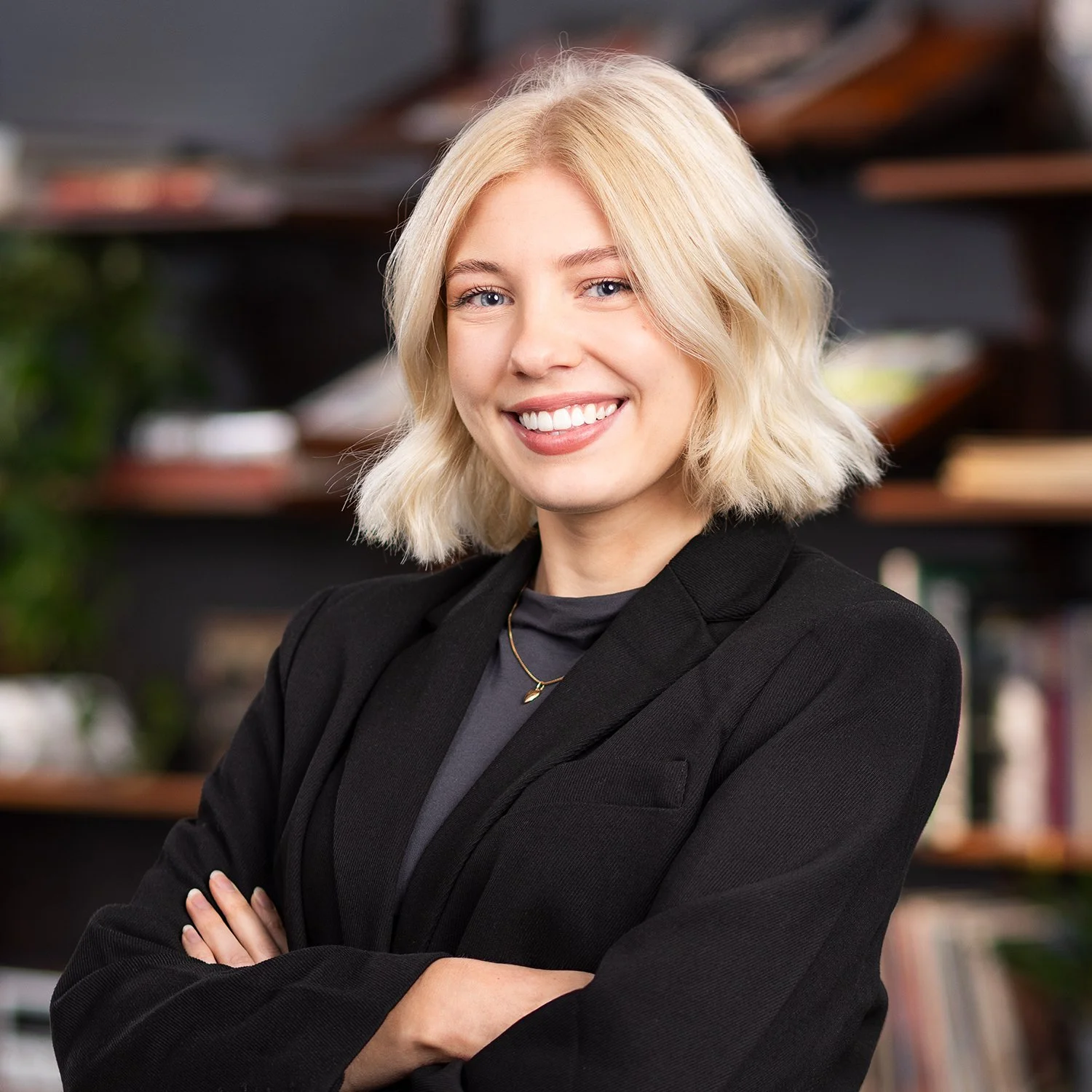 A smiling woman with blonde hair, wearing a black blazer and a necklace, standing with arms crossed in front of a bookcase.