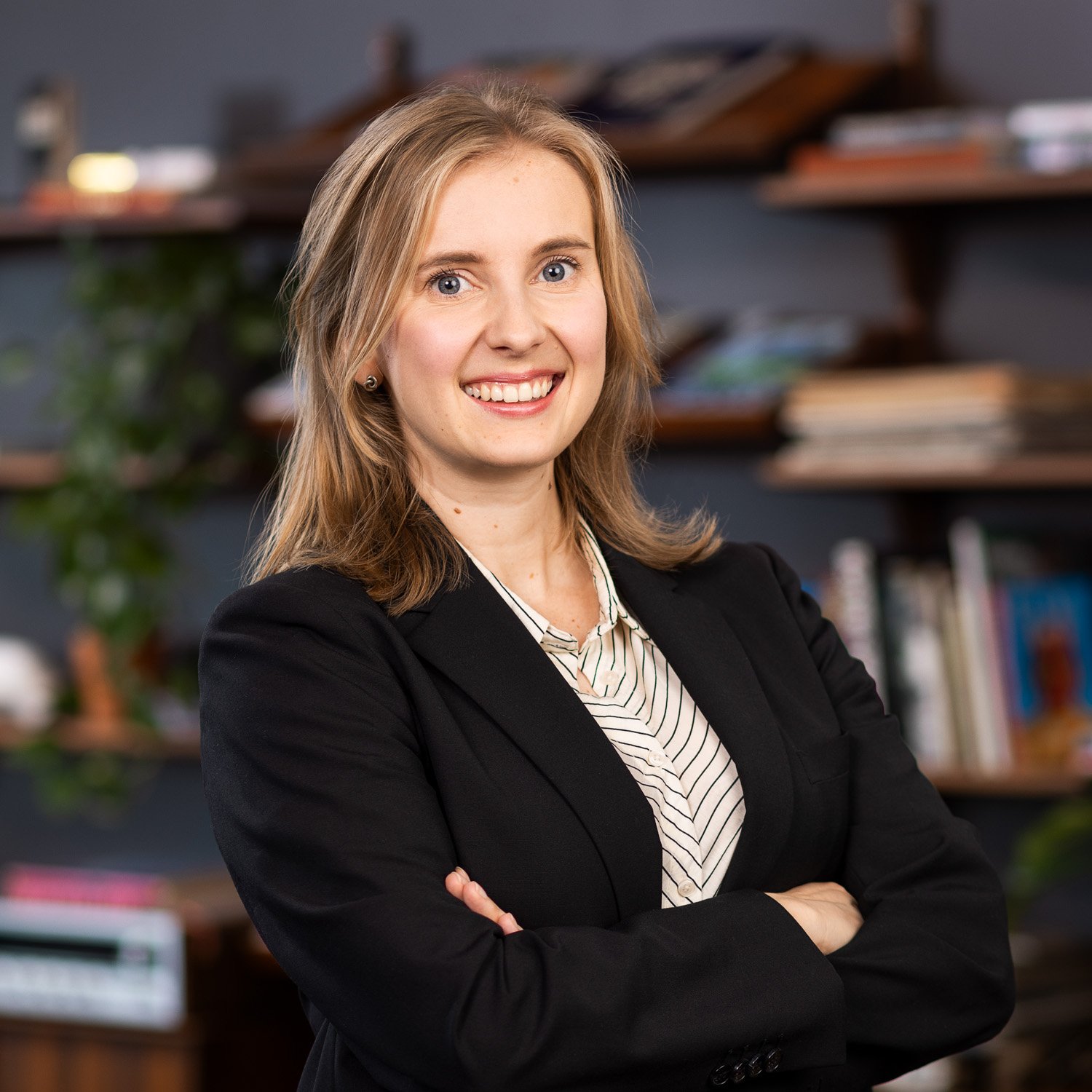 Professional woman with blonde hair and blue eyes, wearing a black blazer and striped shirt, smiling with arms crossed, in an office with shelves of books and decorative items in the background.