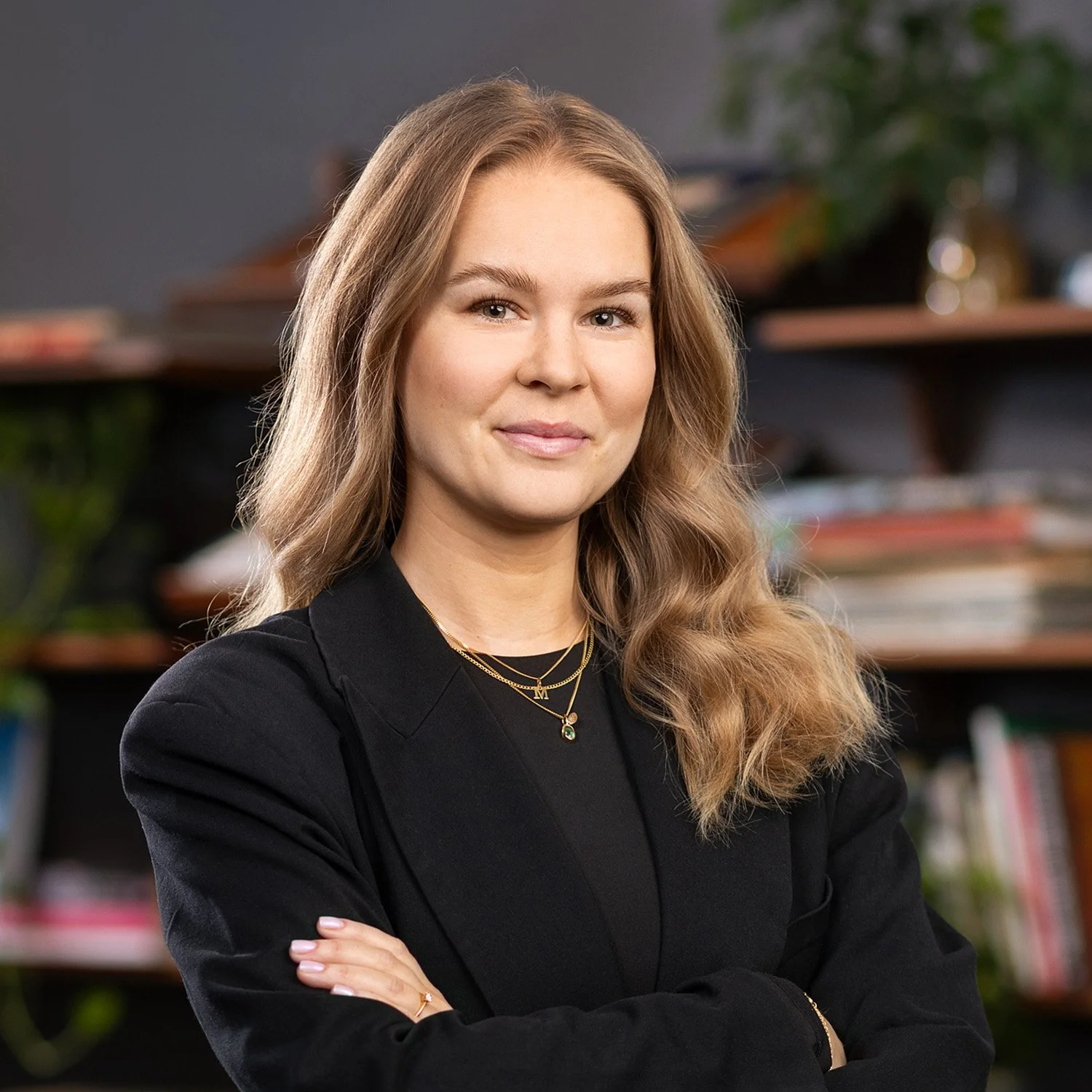 A woman with long wavy blonde hair, dressed in a black blazer and wearing layered necklaces, standing in front of a bookshelf with books and decorative objects.