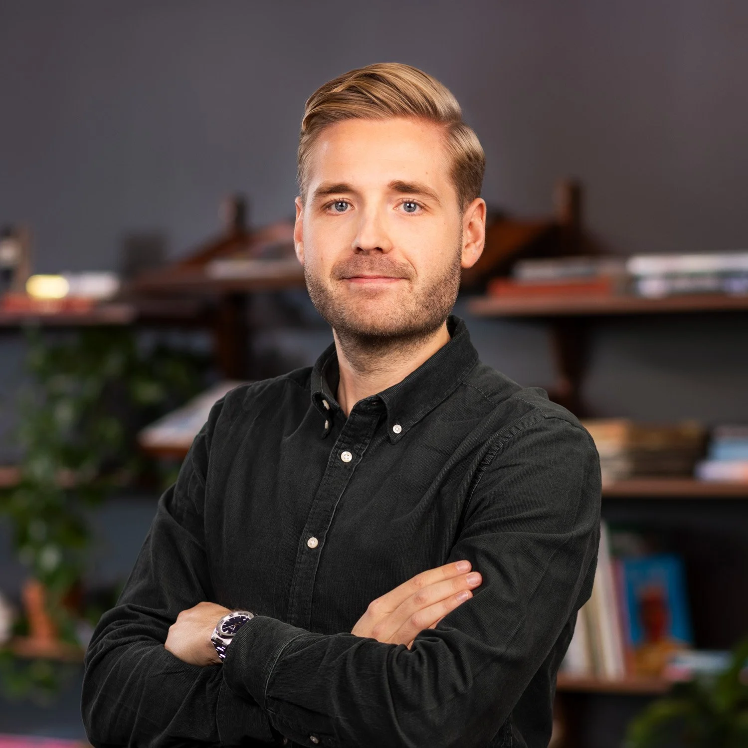 Portrait of a young man with blond hair, beard, wearing a black button-up shirt and a wristwatch, standing with arms crossed in front of a bookshelf.