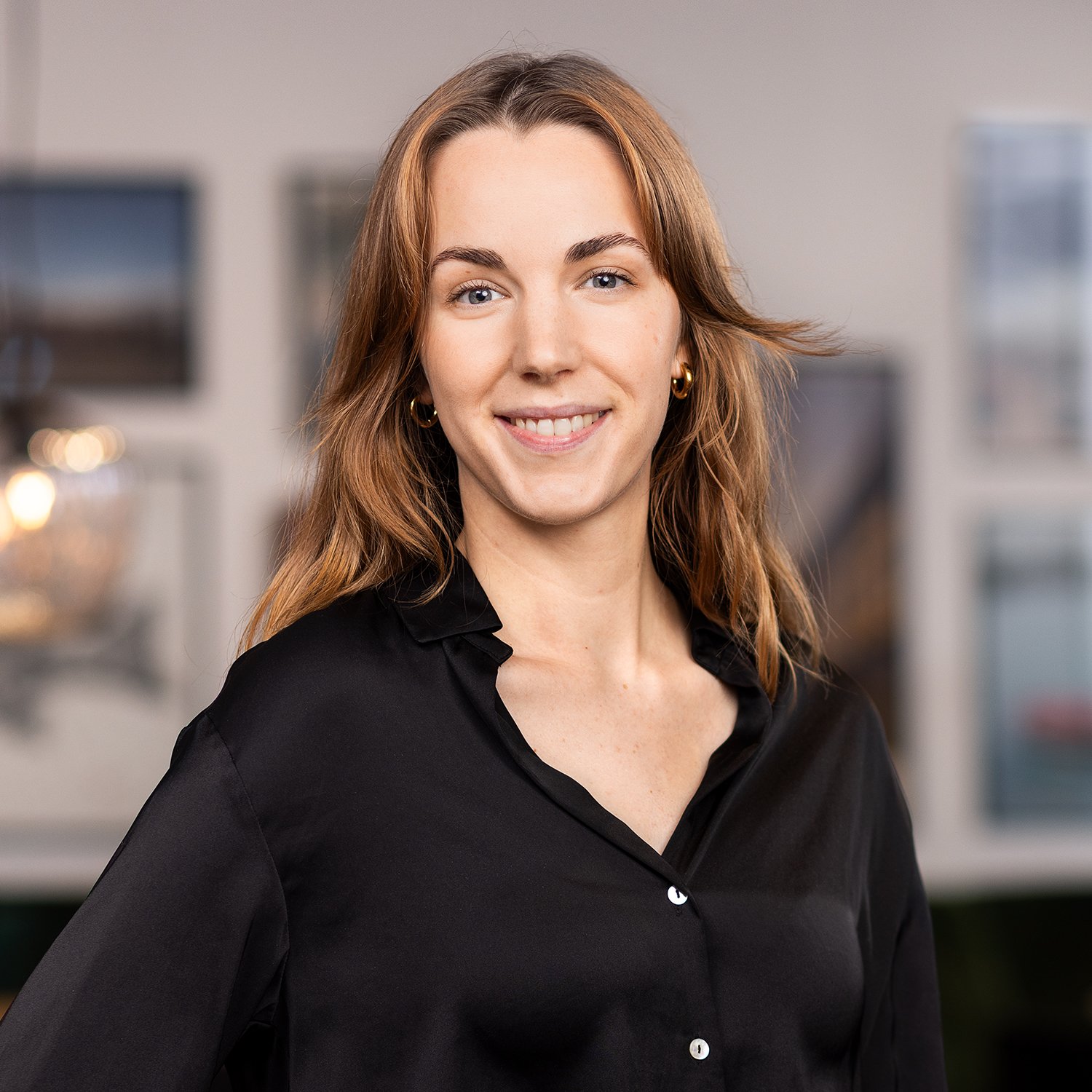 A woman with shoulder-length wavy auburn hair wearing a black button-up shirt, smiling, standing in front of a blurred indoor background.