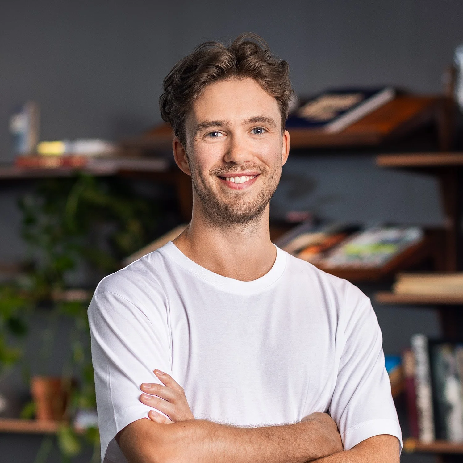 A young man with brown hair and blue eyes, smiling with arms crossed, wearing a white t-shirt, standing indoors with a bookshelf in the background.