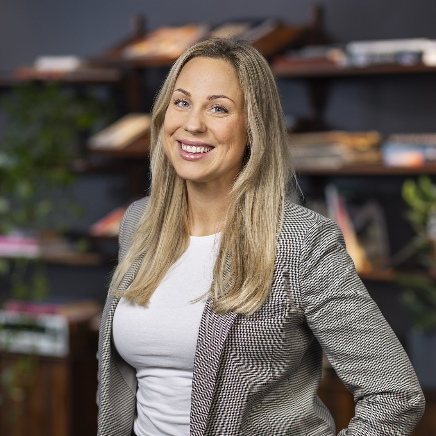 A smiling woman with long blonde hair wearing a checkered blazer and white top standing indoors with bookshelves and a plant in the background.