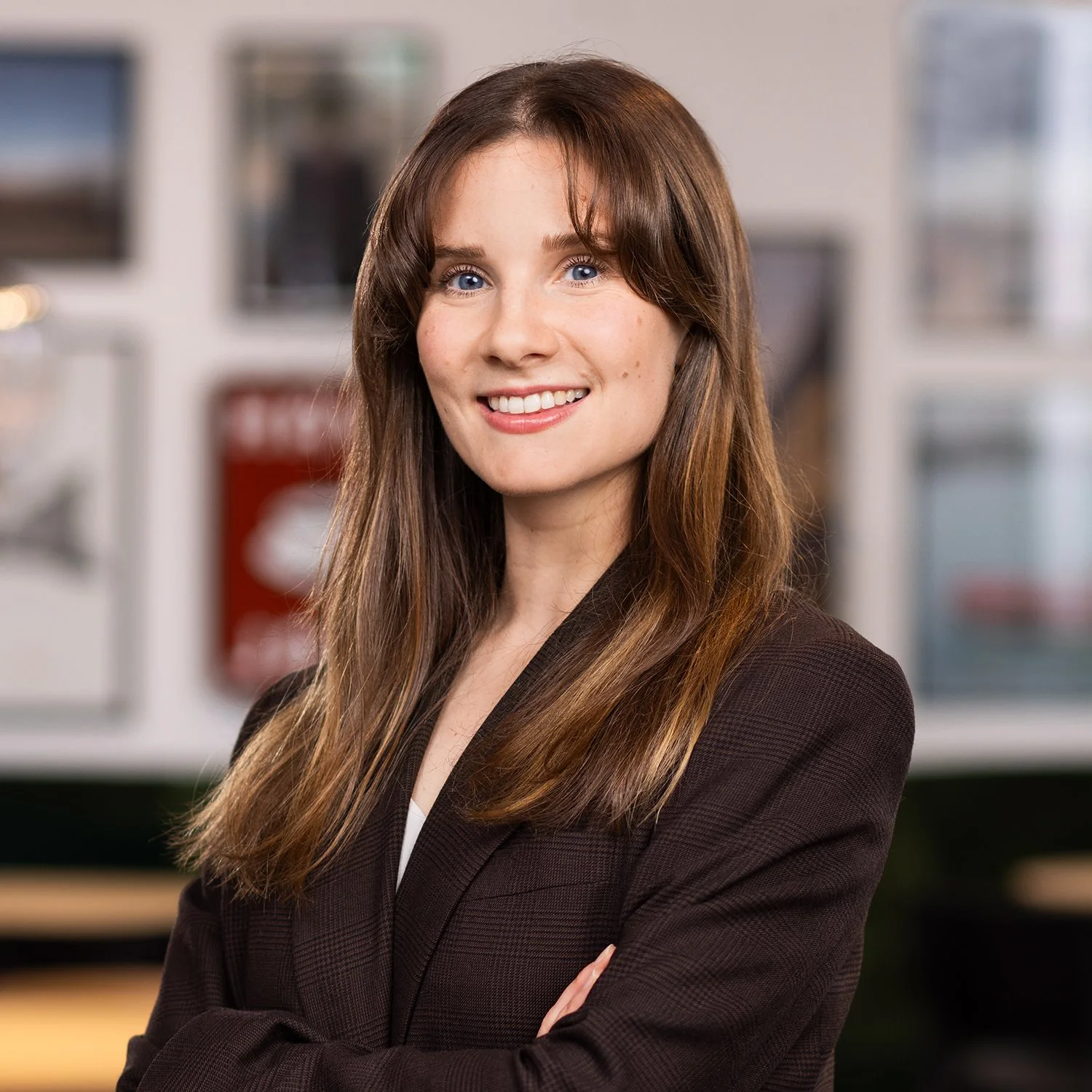 Professional woman with long brown hair in a blazer, smiling confidently with arms crossed, in a modern office environment.