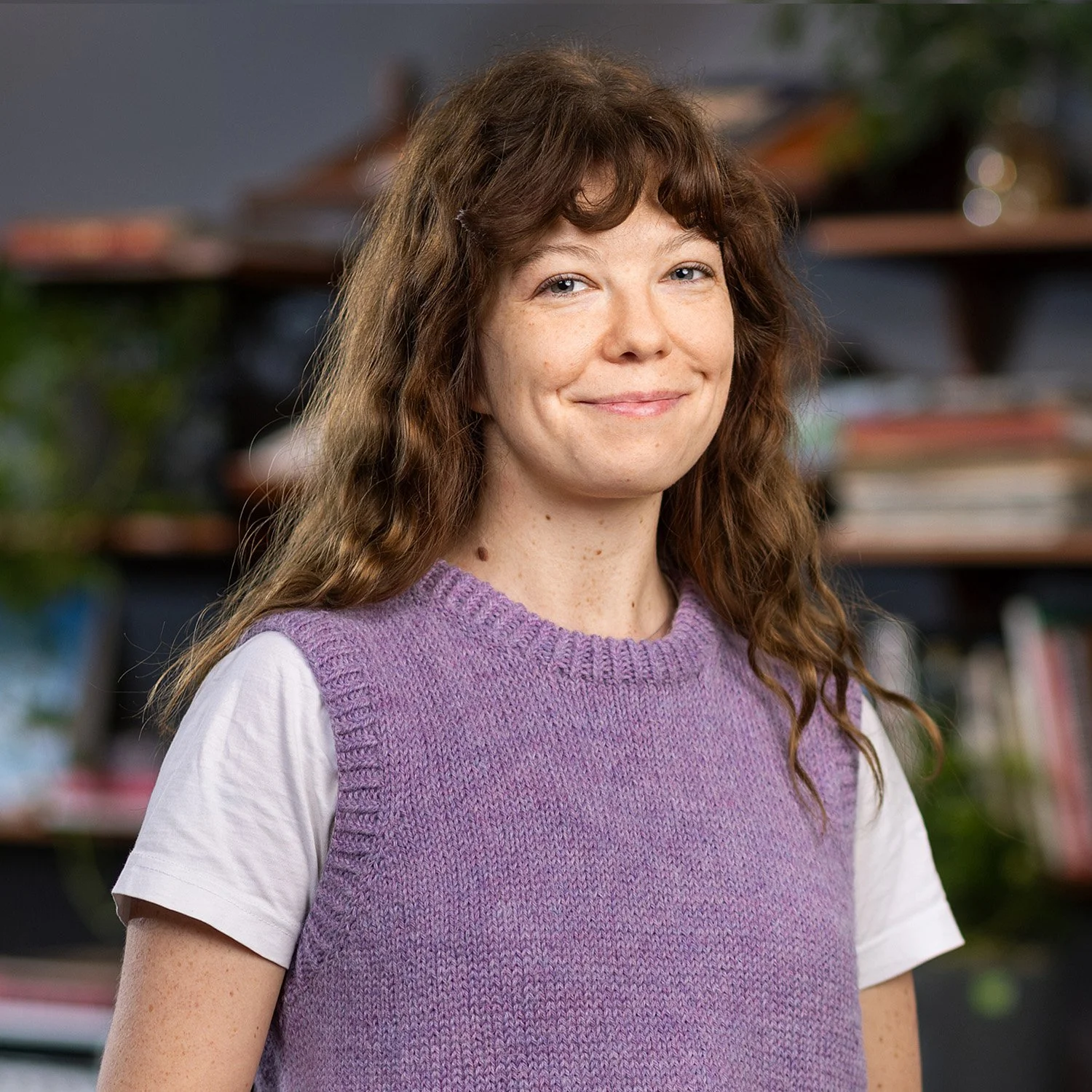 A smiling woman with long curly brown hair wearing a purple knitted vest over a white T-shirt, standing in front of a bookshelf with books and plants.