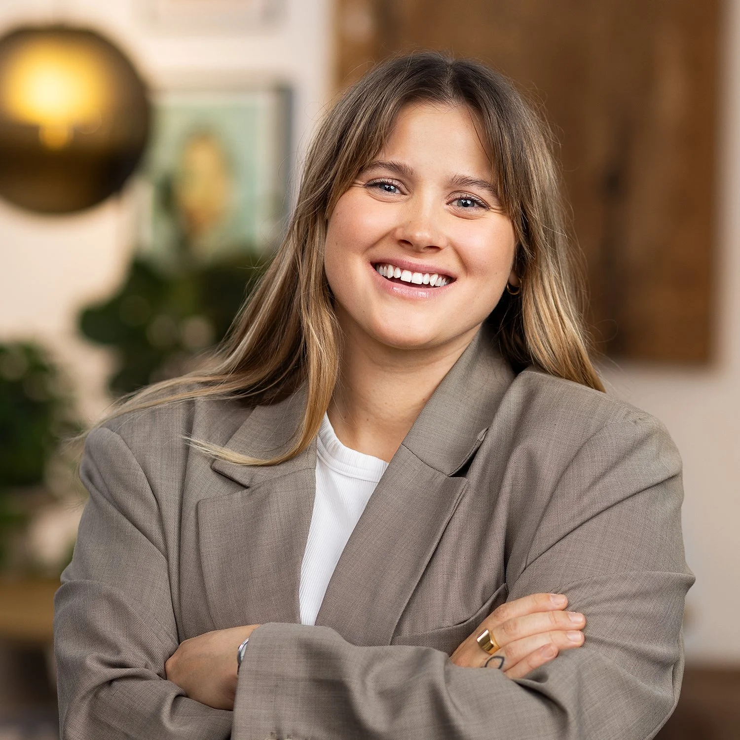 A young woman with long light brown hair smiling with arms crossed, wearing a beige blazer and white shirt, standing in an indoor space with blurred background.