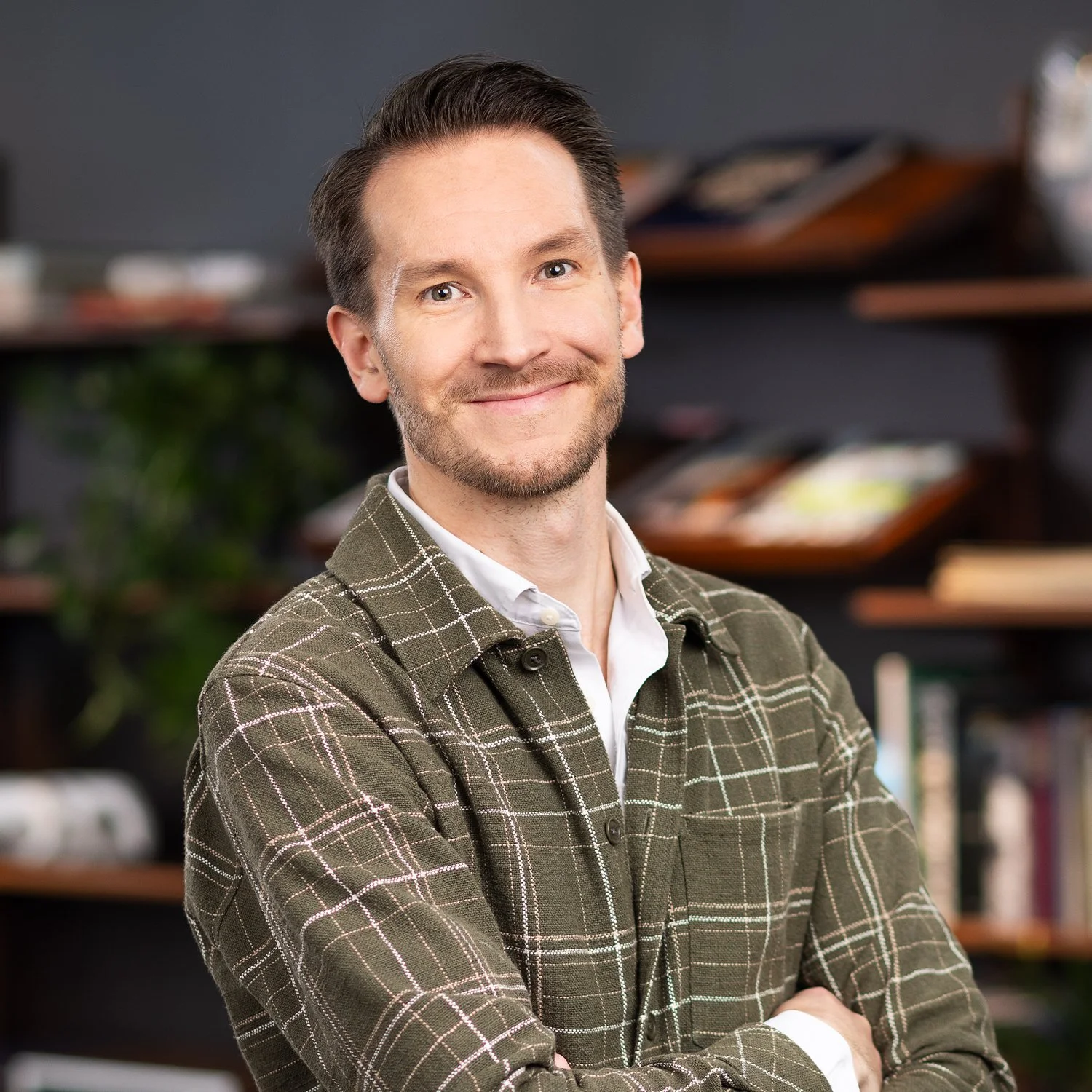 A man with short dark hair and a beard smiling at the camera, wearing a green plaid shirt over a white collared shirt, standing in front of dark shelves filled with books and decorative items.