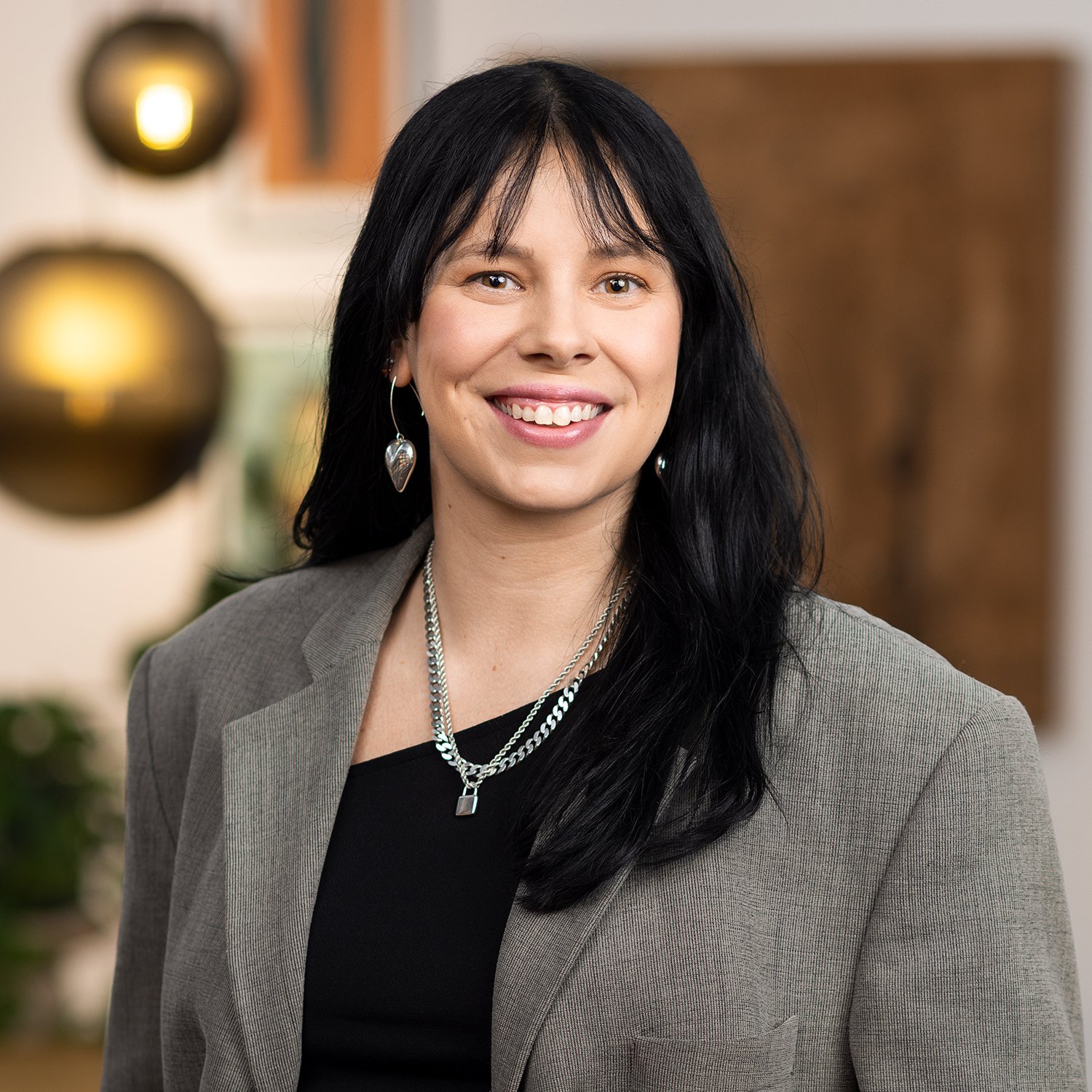 A woman with long black hair, wearing a gray blazer, black top, silver jewelry, and smiling, standing indoors with a blurred background.