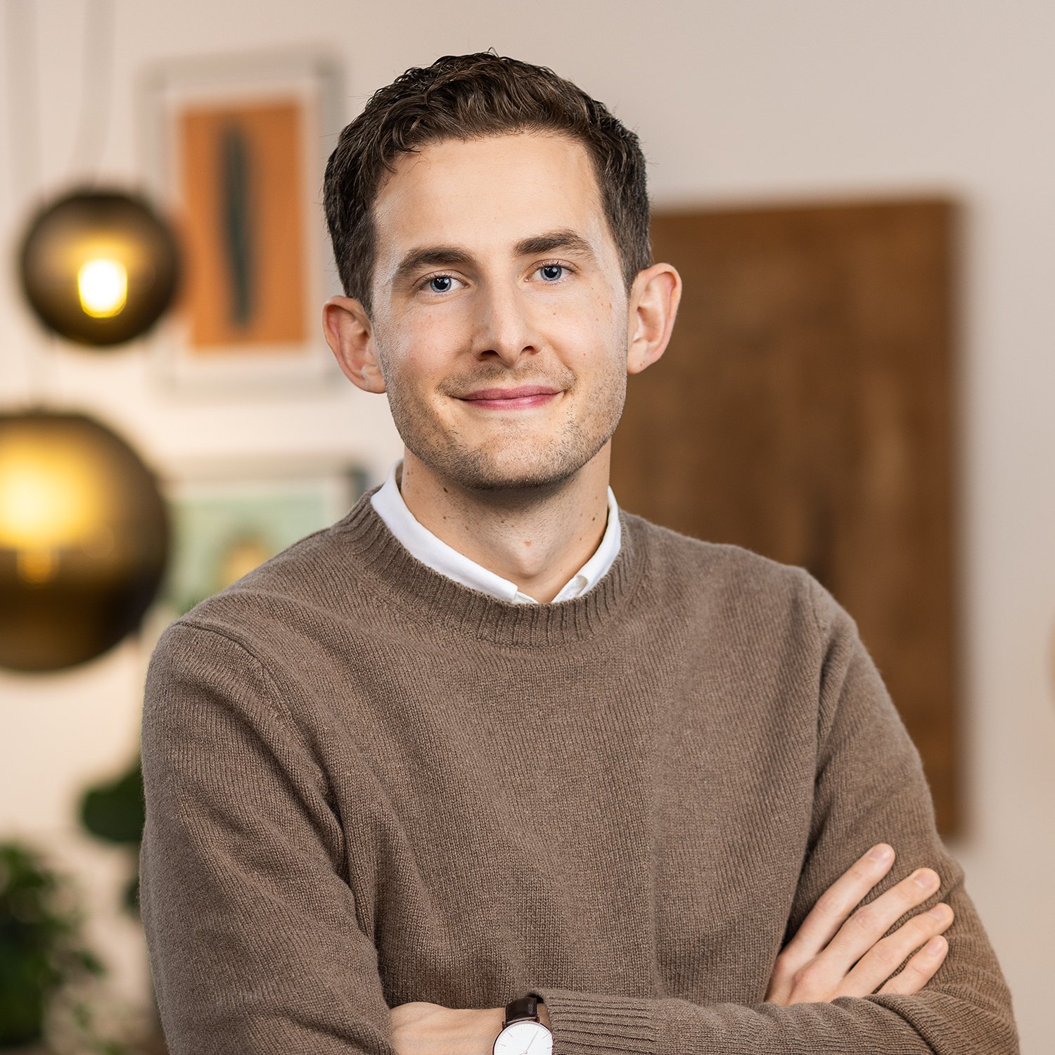 A young man with dark hair and blue eyes, wearing a brown sweater over a white collared shirt, standing with his arms crossed in a warmly lit indoor setting.