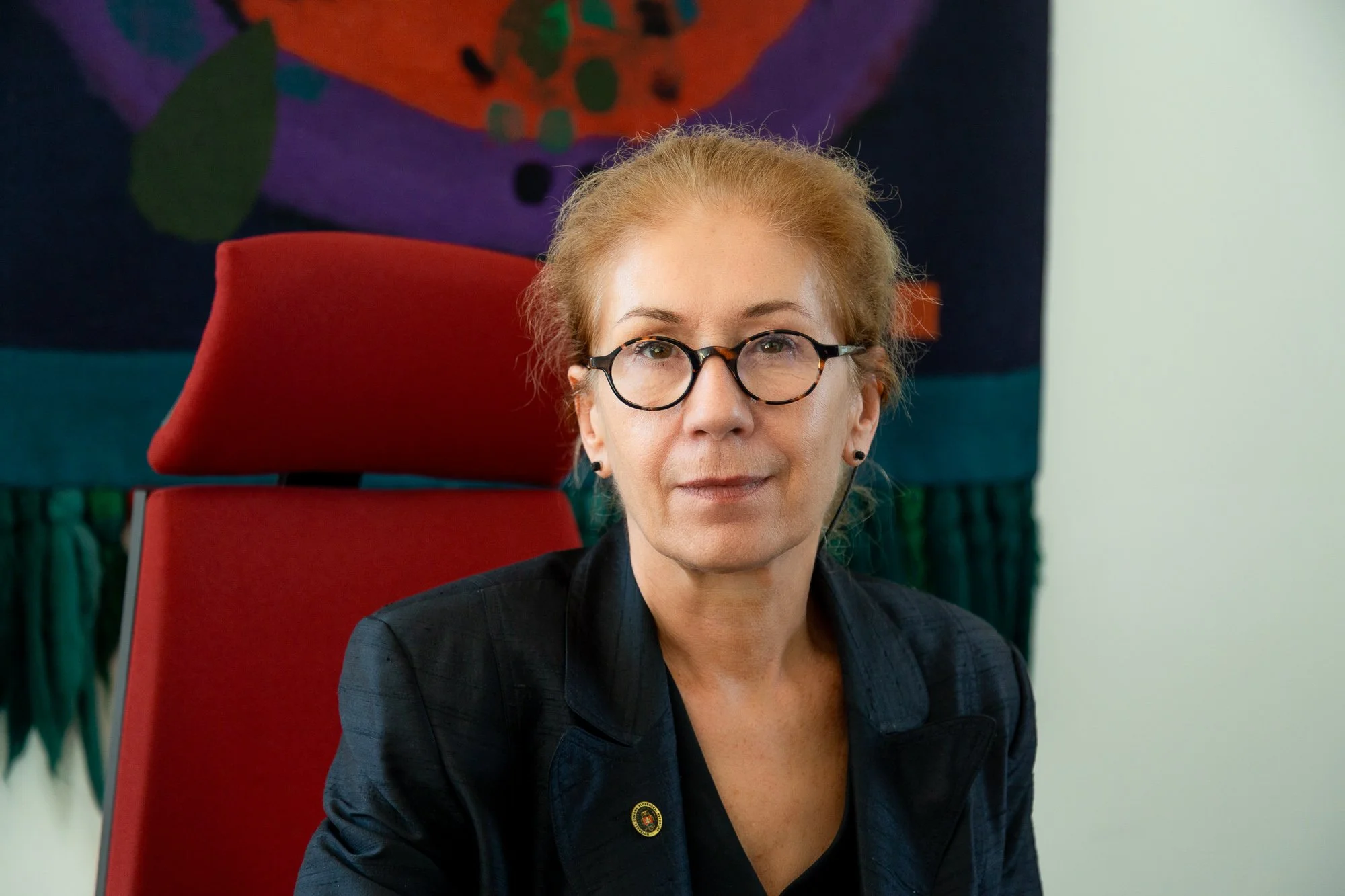 Notárka stredného veku s okuliarmi a svetlými vlasmi, sediaca na červenej stoličke pred farebným tapisériovým pozadím.