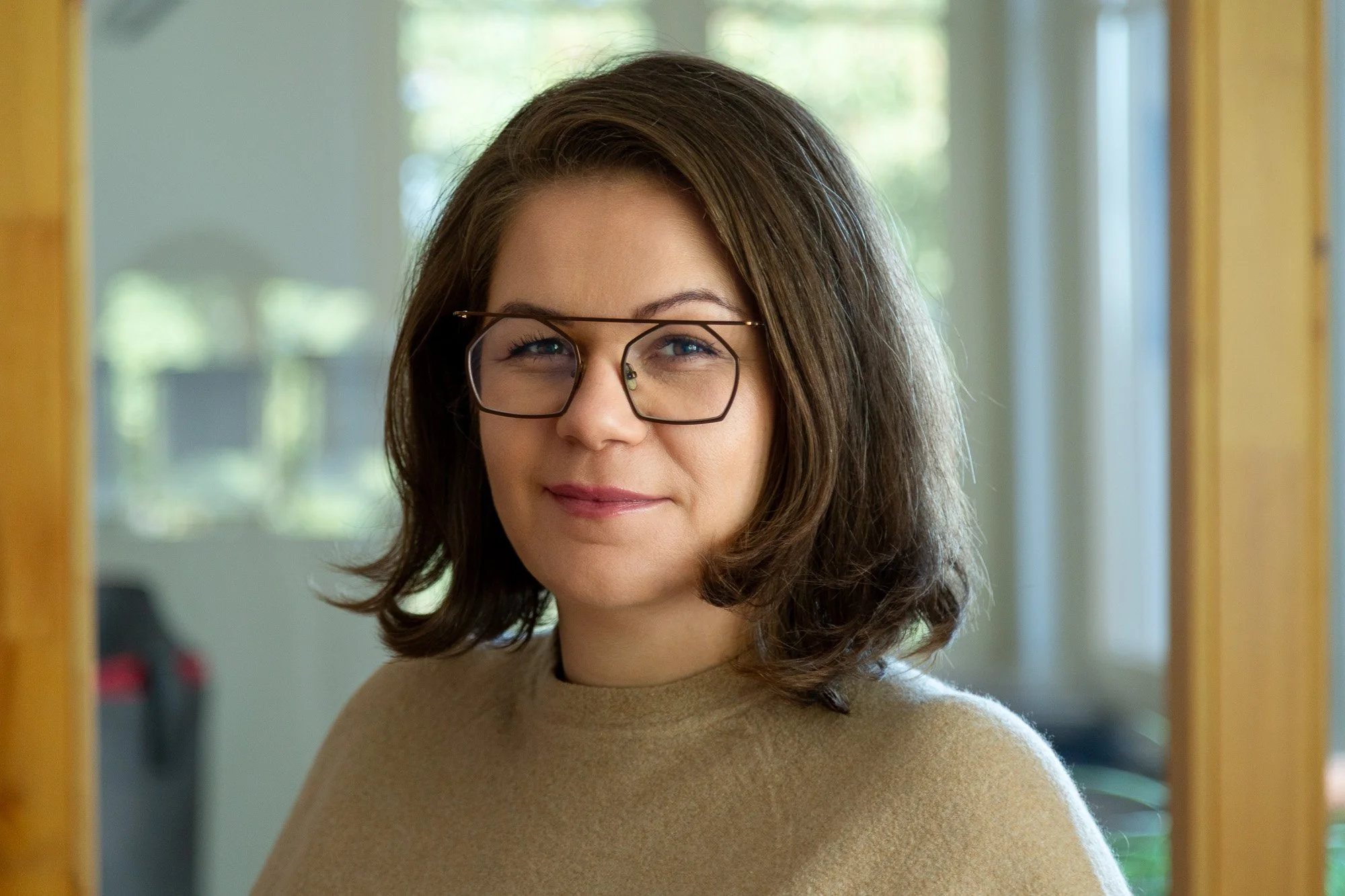 Notárka so stredne dlhými hnedými vlasmi a okuliarmi, usmievajúca sa v kancelárii s oknami a zelenými rastlinami v pozadí.