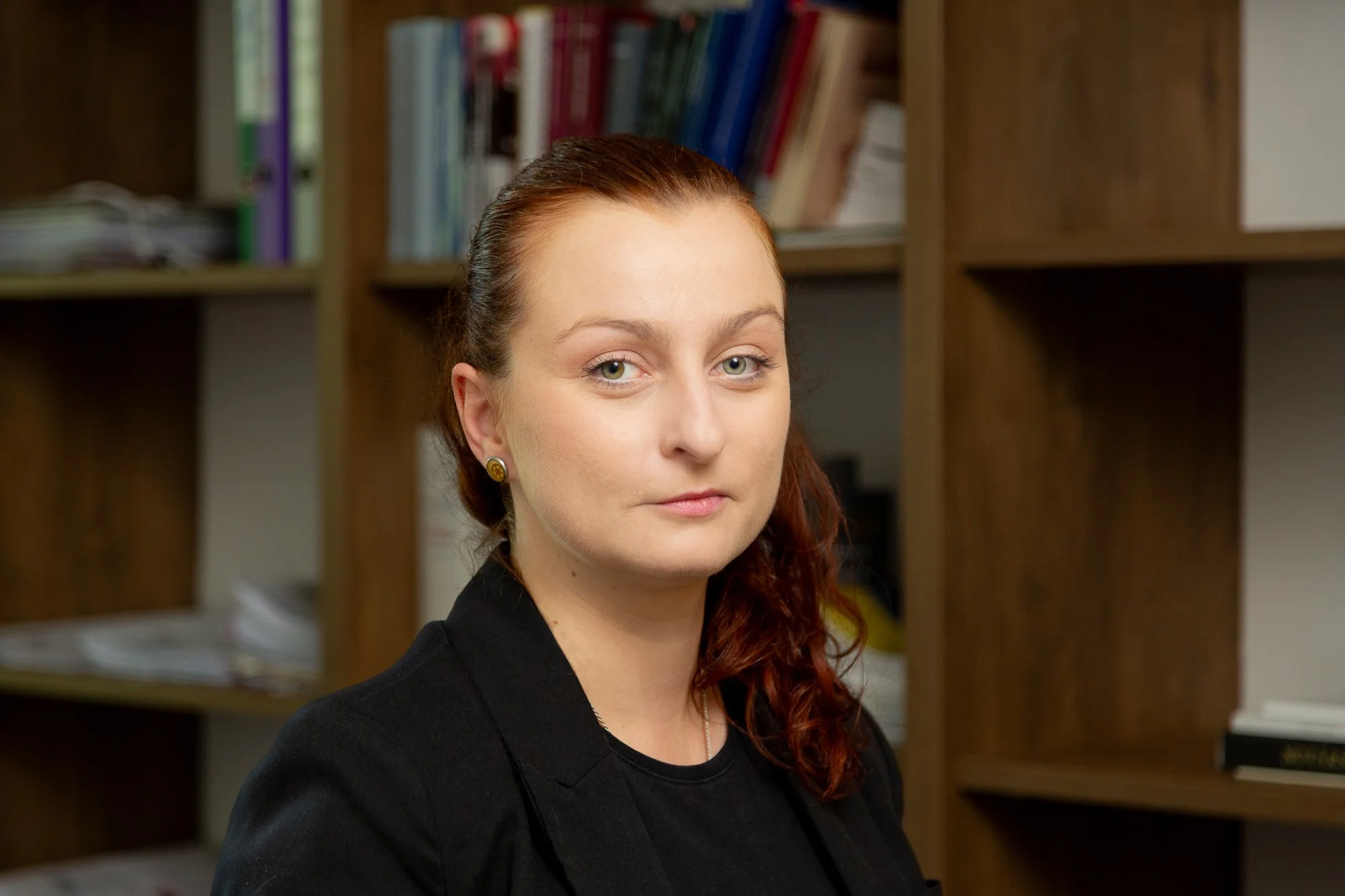 Notárka s červenými vlasmi v čiernom saku a náušnicami stojaca pred drevenými knižnicami plnými kníh a dokumentov.