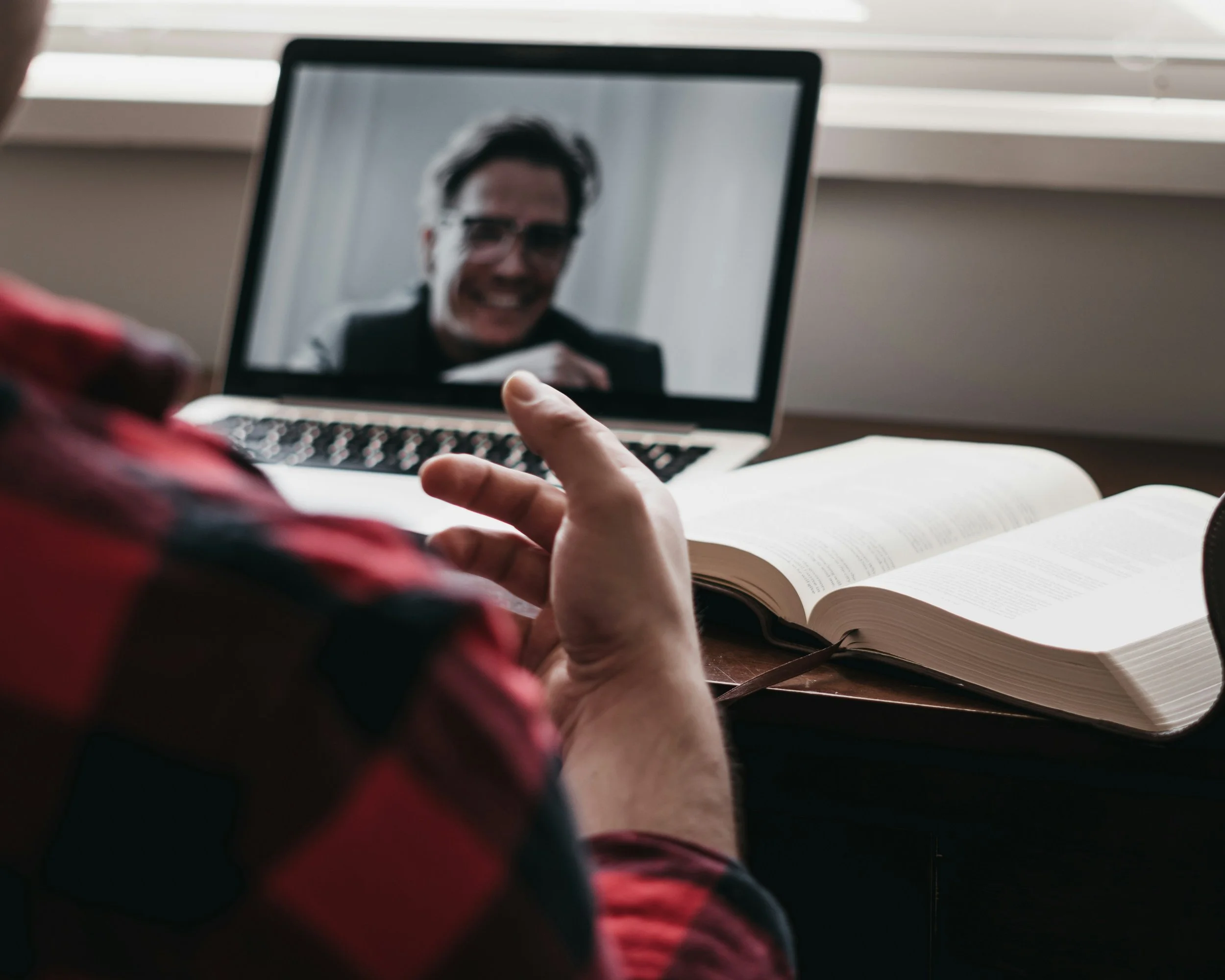 Person on a video call with a man on a laptop screen. Open book and notebook on the desk.