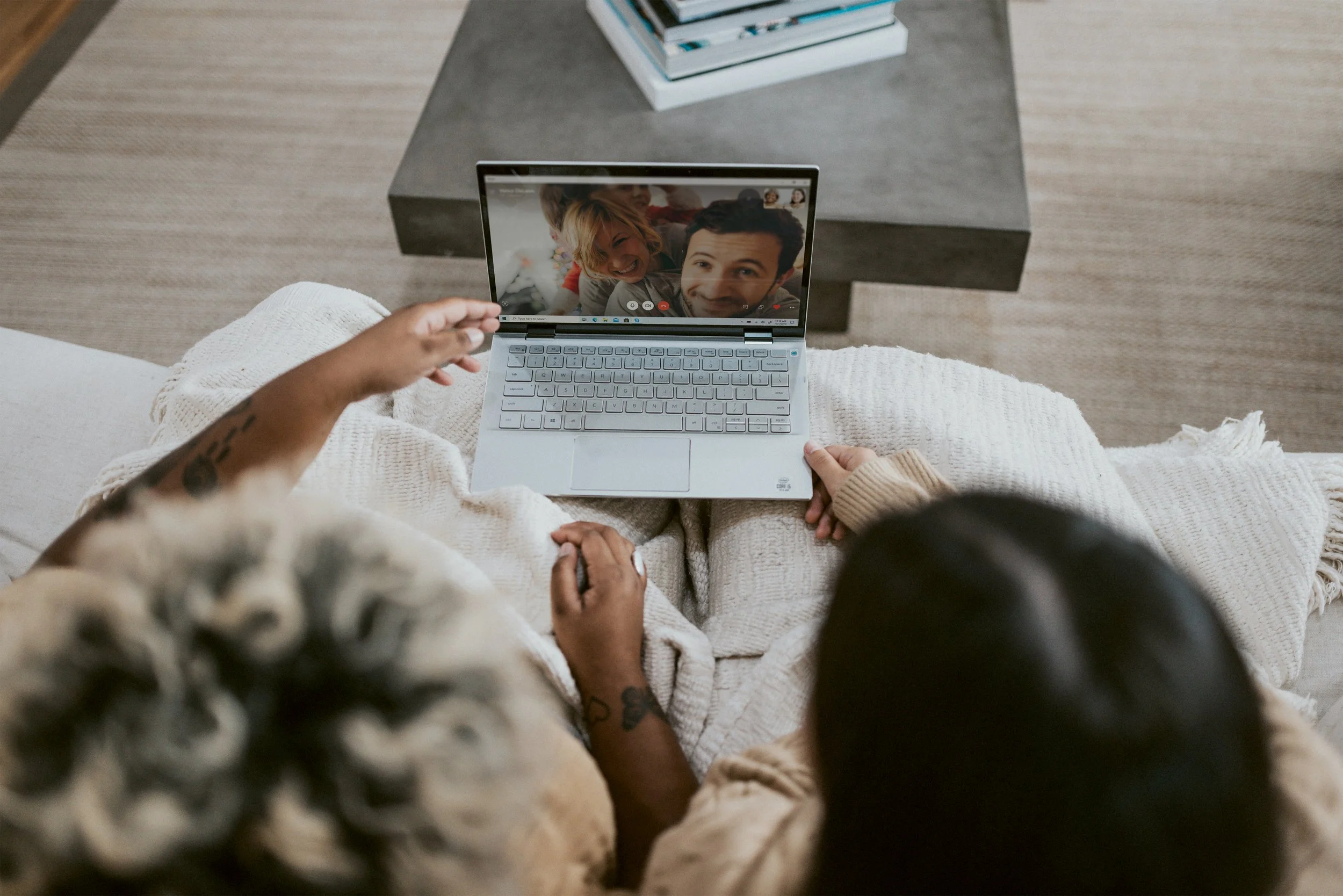 A person sitting on a couch holding a laptop, video chatting with two smiling children. The scene is viewed from above, with a coffee table and books in the background.