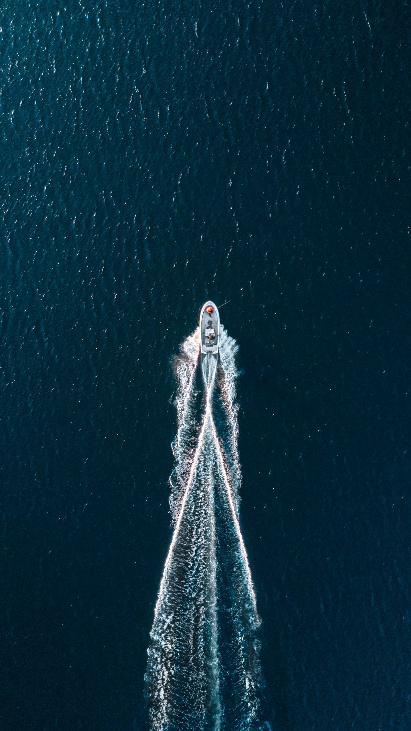 An aerial view of a boat moving swiftly across dark blue water, creating white foamy wakes behind it.