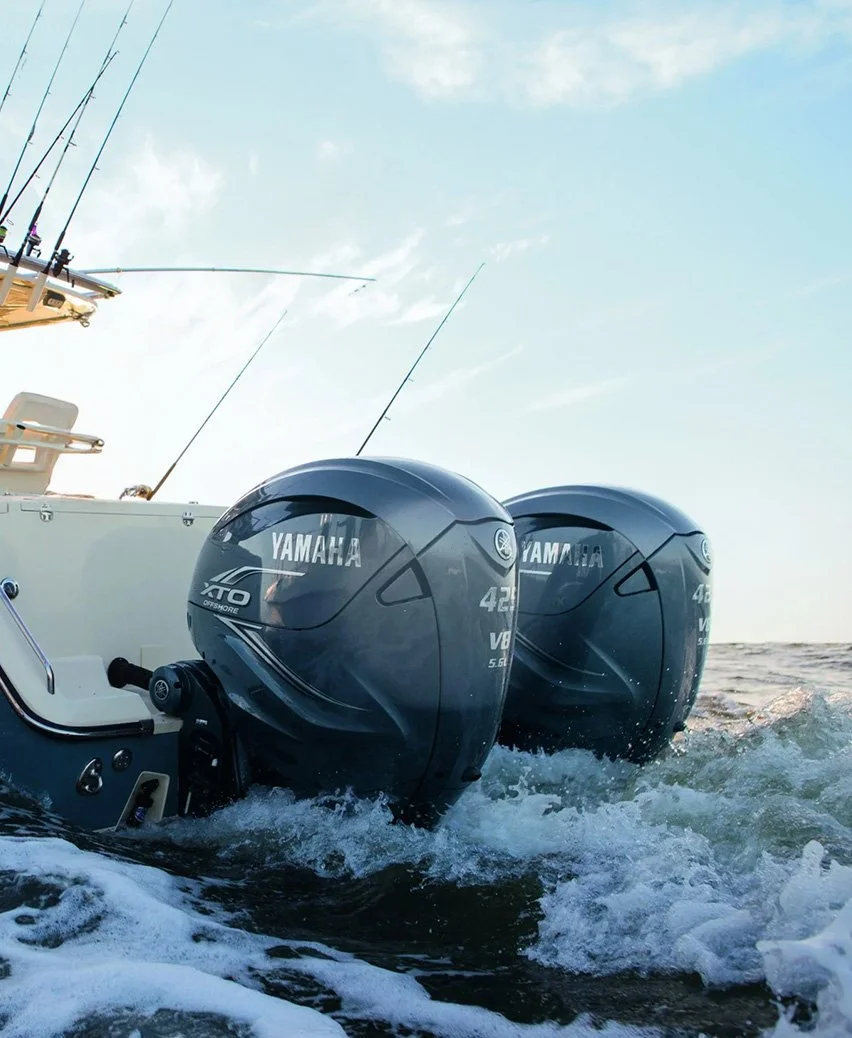 Two Yamaha outboard motors attached to a boat, with waves splashing around them on the water. The sky is partly cloudy.