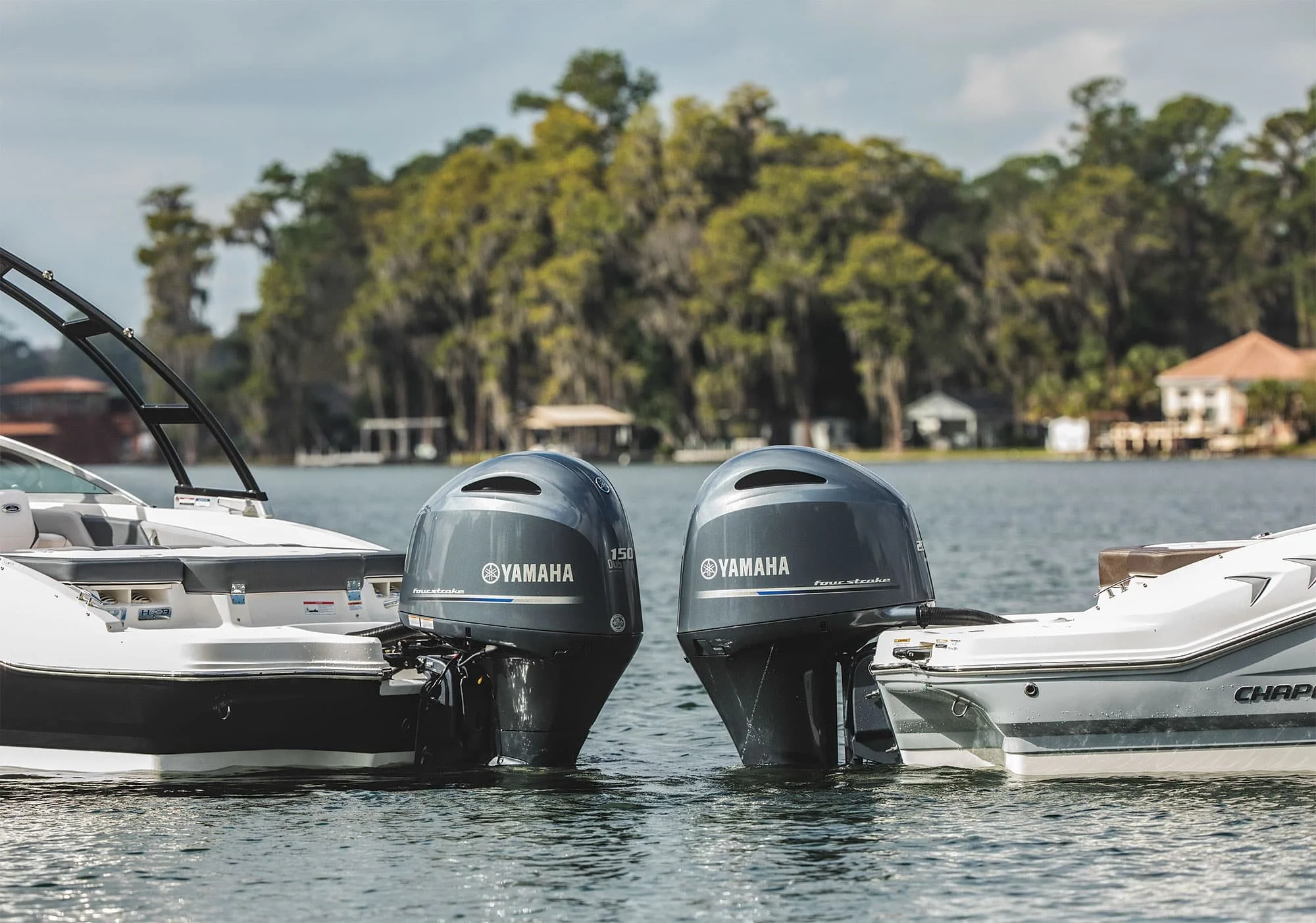 Two Yamaha outboard motors mounted on a boat near the water with trees and houses in the background.