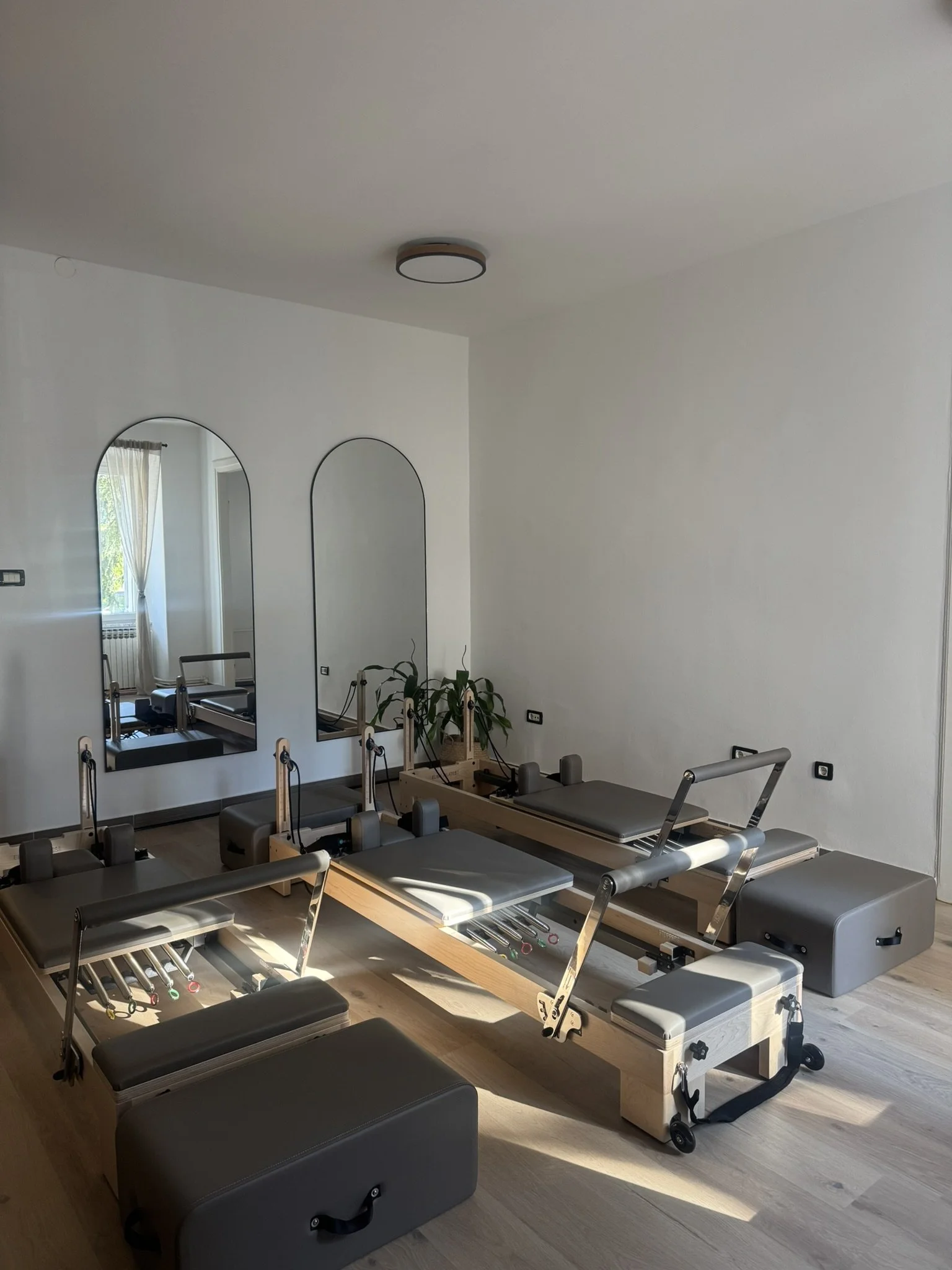 Pilates reformer equipment in a bright, minimalistic studio room with wooden floors, two large mirrors on the wall, and a window with curtains.  in a premium reformer pilates studio in Rijeka, Croatia