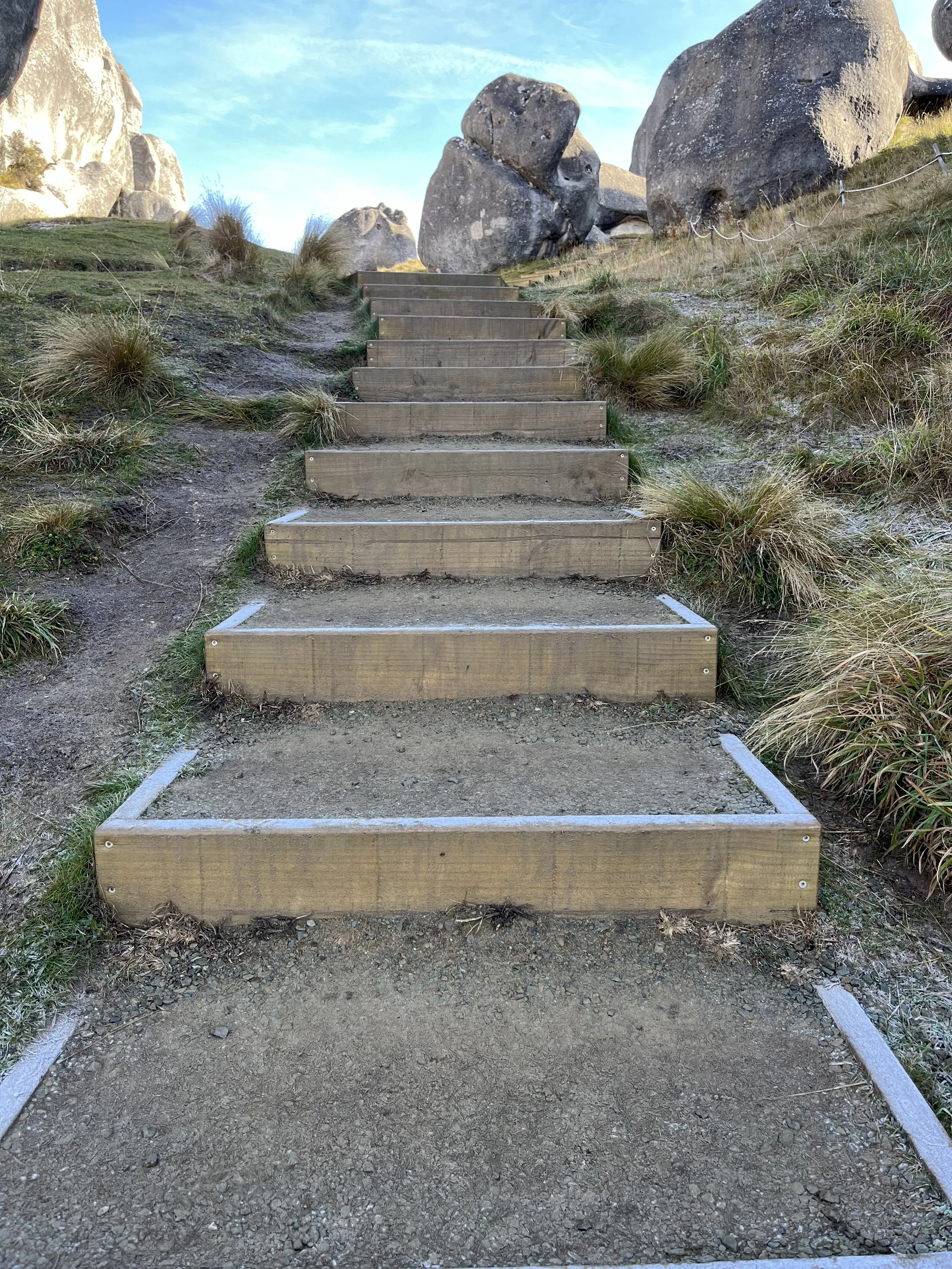 Wooden stairs on a dirt path leading up a grassy hillside with large boulders at the top and a blue sky with some clouds in the background.