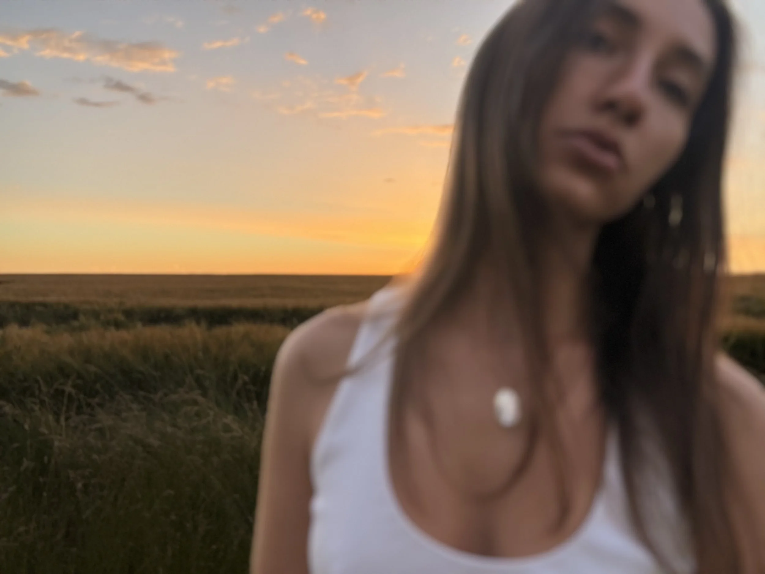 Jeune femme aux cheveux longs portant un haut blanc, debout dans un champ lors d'un coucher de soleil avec un ciel orangé et quelques nuages.