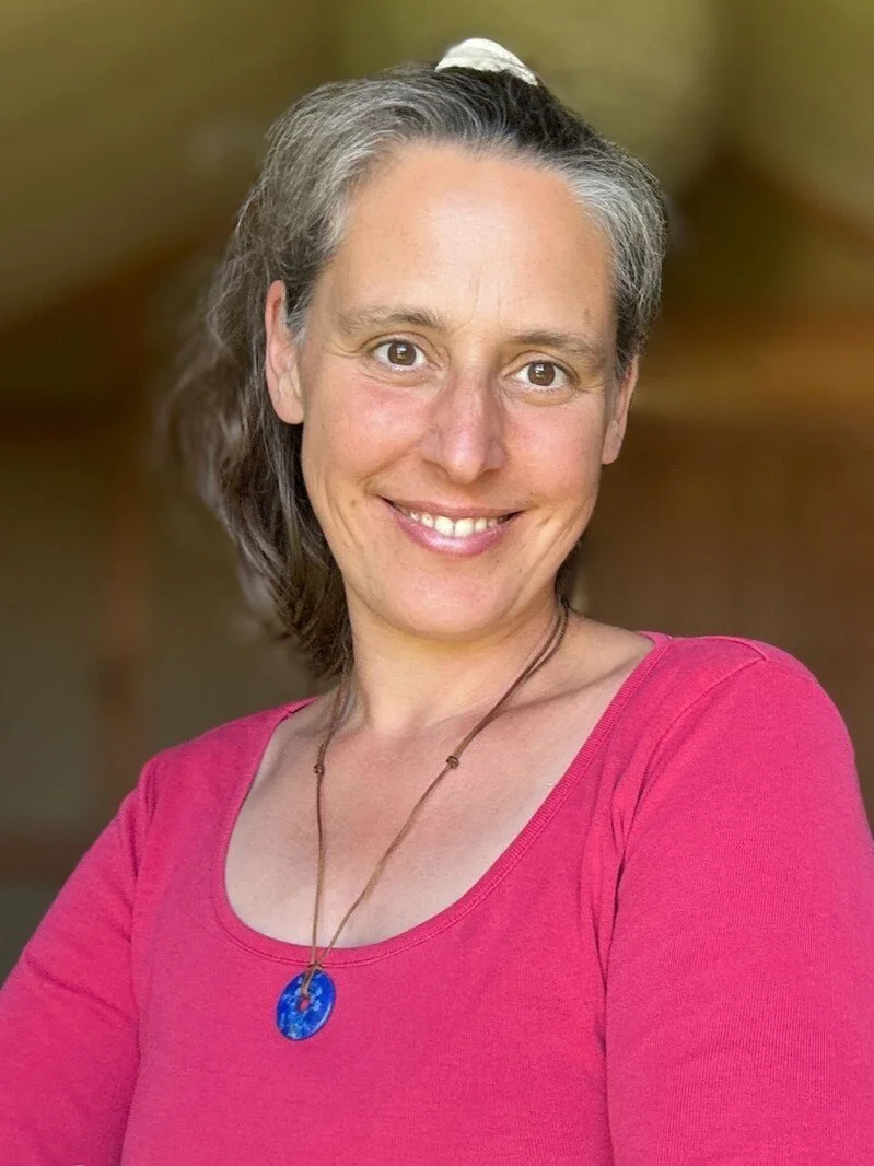 Porträt einer lächelnden Frau mit grau-braunen Haaren, trägt eine rosa Bluse und eine blaue Halskette.