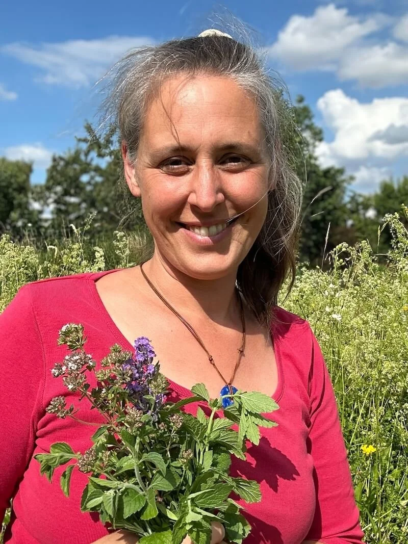 Eine lachende Frau mit braunem Haar in einem roten Shirt, die in einem blühenden Feld mit grünen Pflanzen steht, hält einen Strauß frischer Kräuter, während sie an einem sonnigen Tag im Freien steht.