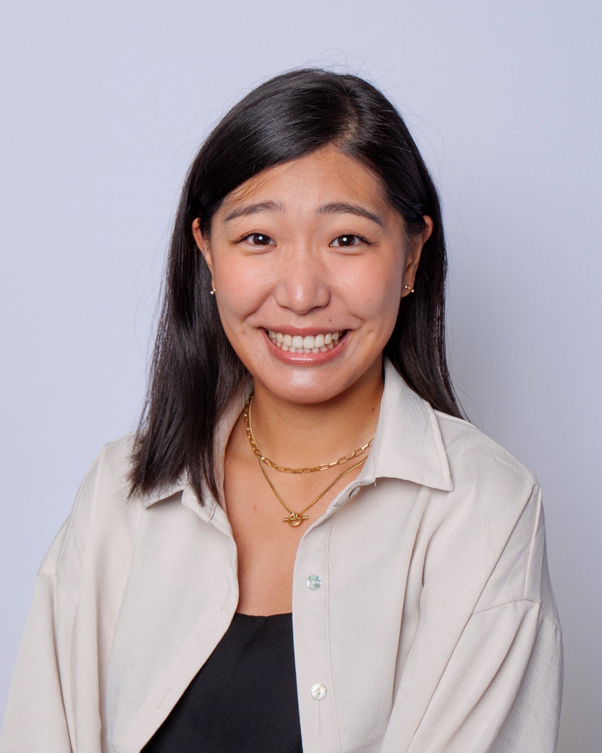Smiling woman with dark hair wearing a beige shirt and layered gold necklaces against a light grey background.