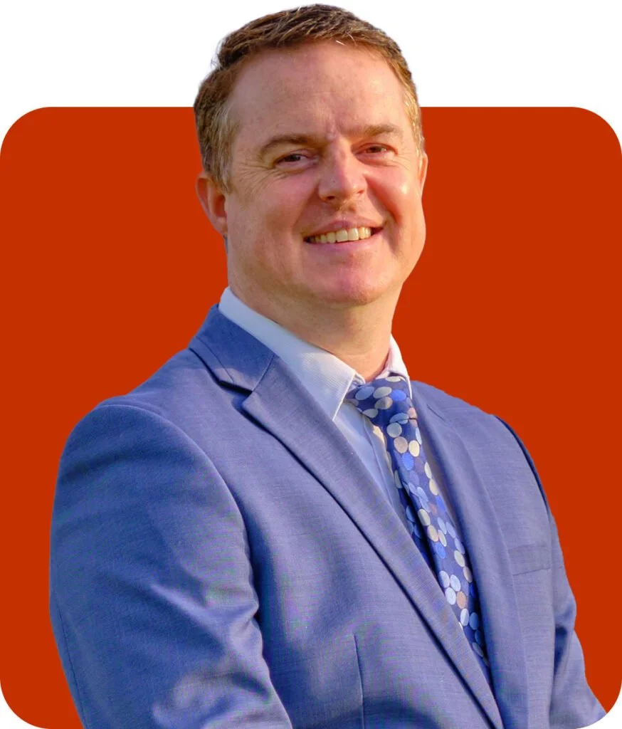 Portrait of a smiling man in a blue suit and patterned tie against a red background.